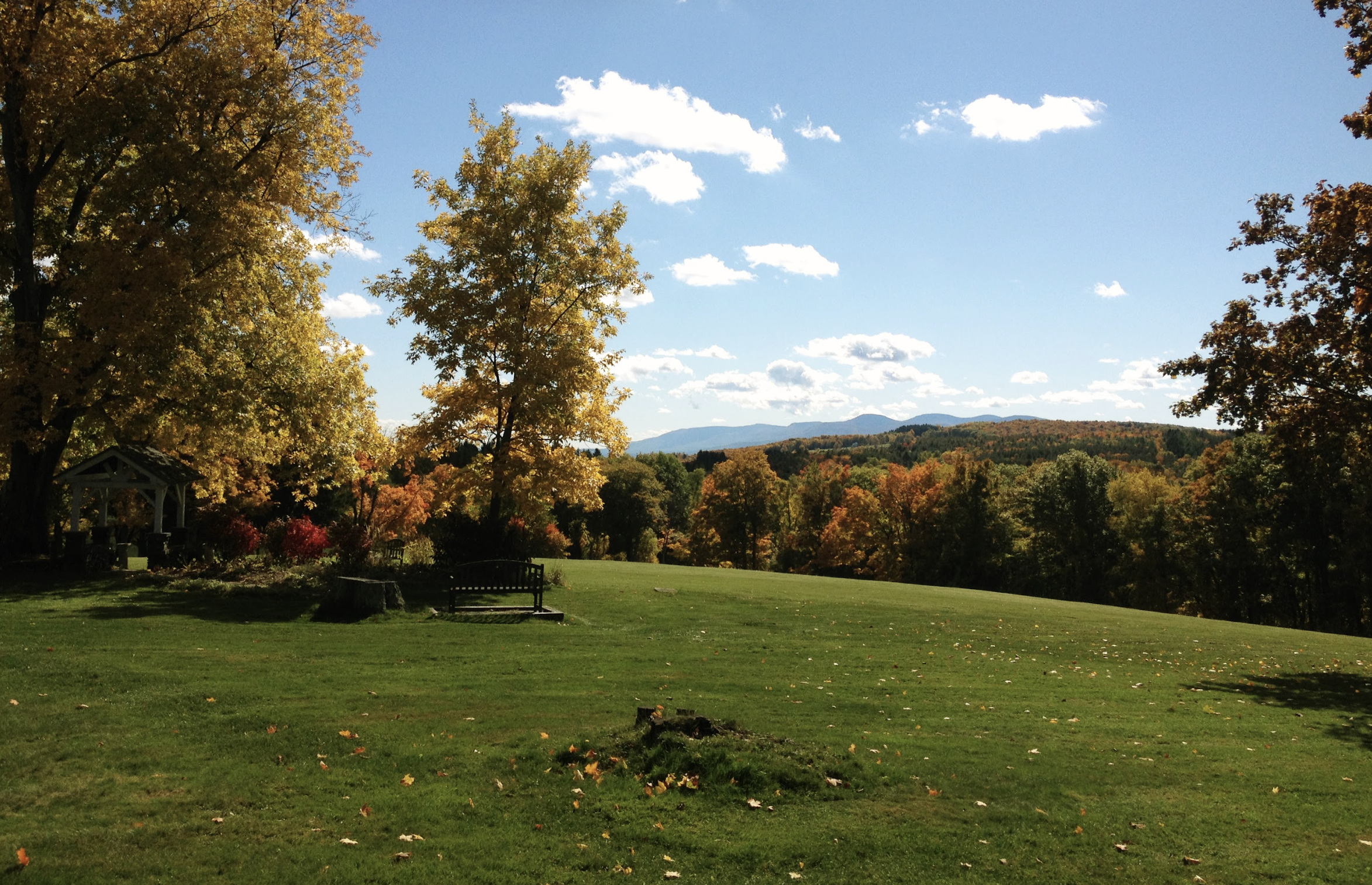 A scenic park scene with green grass, colorful autumn trees in yellows, oranges, and reds, a bench, a garden gazebo, and mountains in the background under a bright blue sky with scattered white clouds.