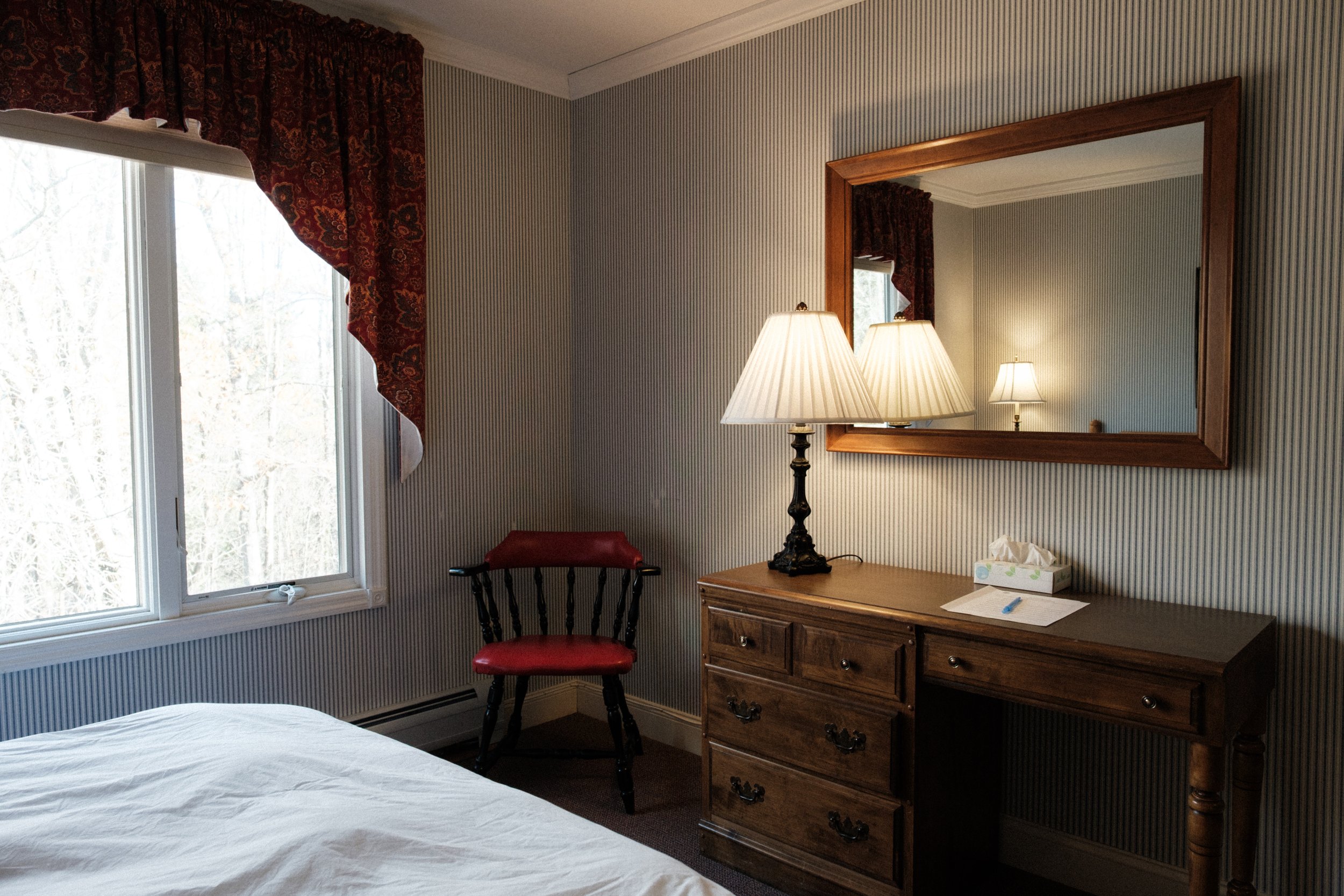 A bedroom corner with a window, red curtain, wooden dresser with a lamp, mirror, tissue box, and a red and black chair.