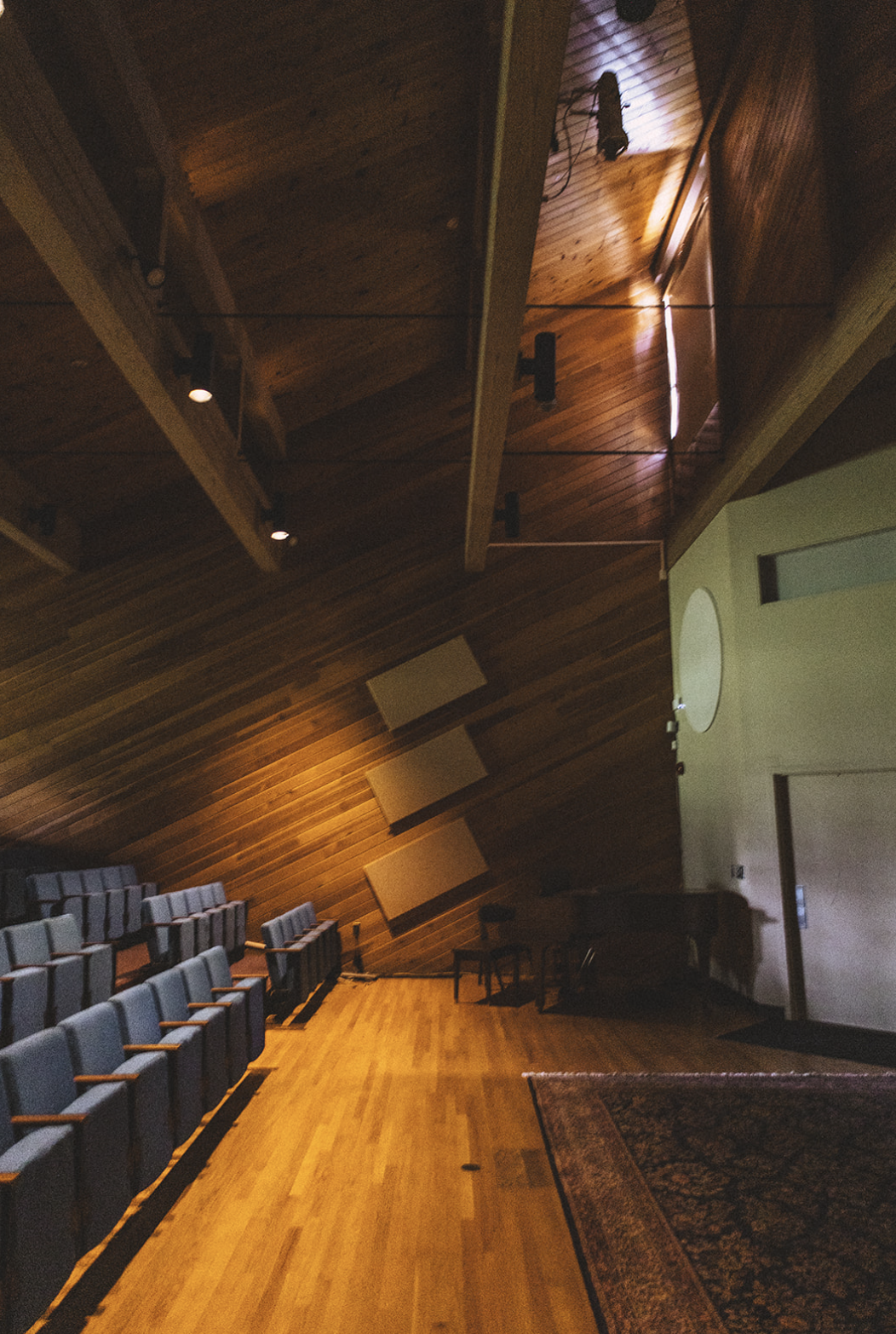 Empty auditorium with rows of blue seats, wooden walls and ceiling, a rug on the floor, and a white circular object on the wall.