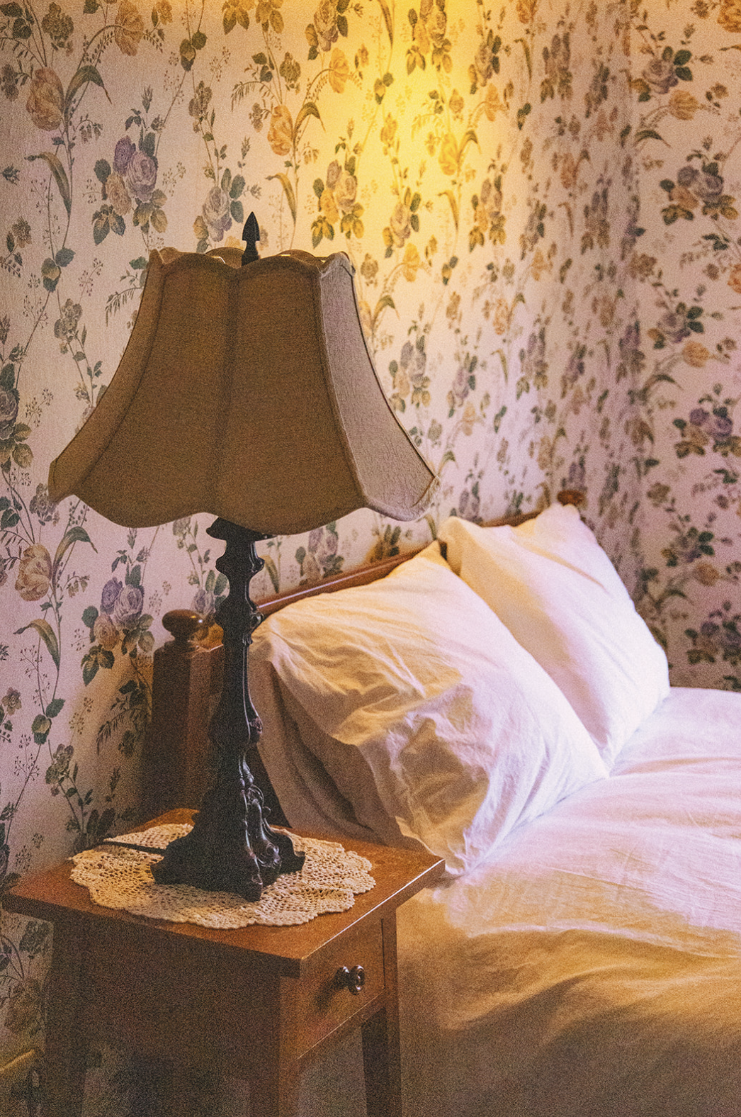A vintage bedroom corner featuring a bed with white pillows and bedding, a wooden nightstand with a lace doily, and a traditional table lamp with a fabric shade, set against floral wallpaper.