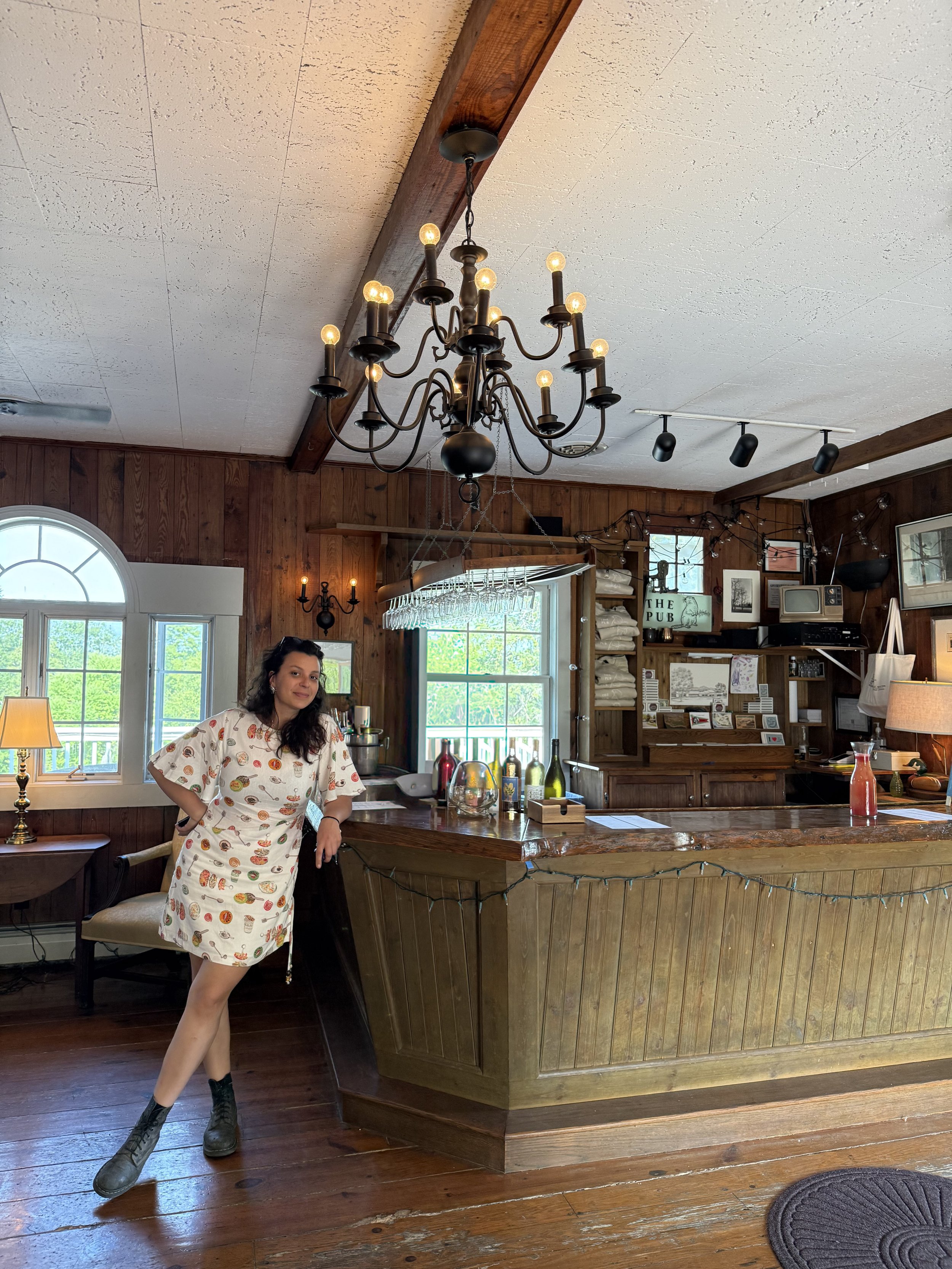 A woman standing inside a cozy, rustic bar or pub with wooden walls and floors, sitting near a wooden bar counter decorated with bottles and glassware. There are windows, a chandelier, and various decorations in the background.