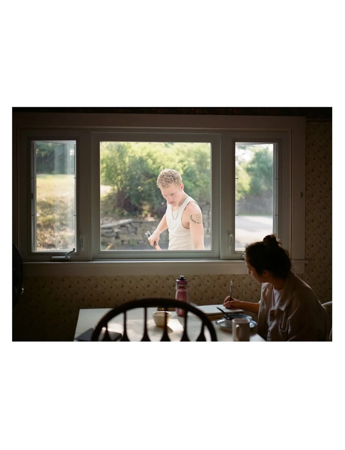 A woman sitting at a kitchen table writing, with a window through which a young man is visible outside, looking down and holding something in his hand.
