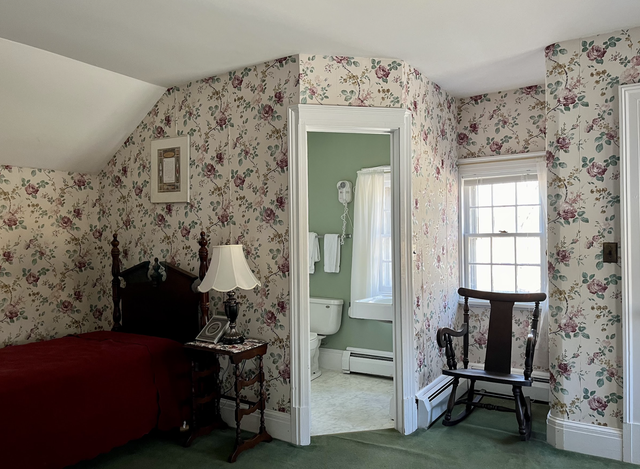 Bedroom with floral wallpaper, a wooden bed, nightstand with lamp, a rocking chair, and a window opening to a small bathroom with green walls and a toilet.
