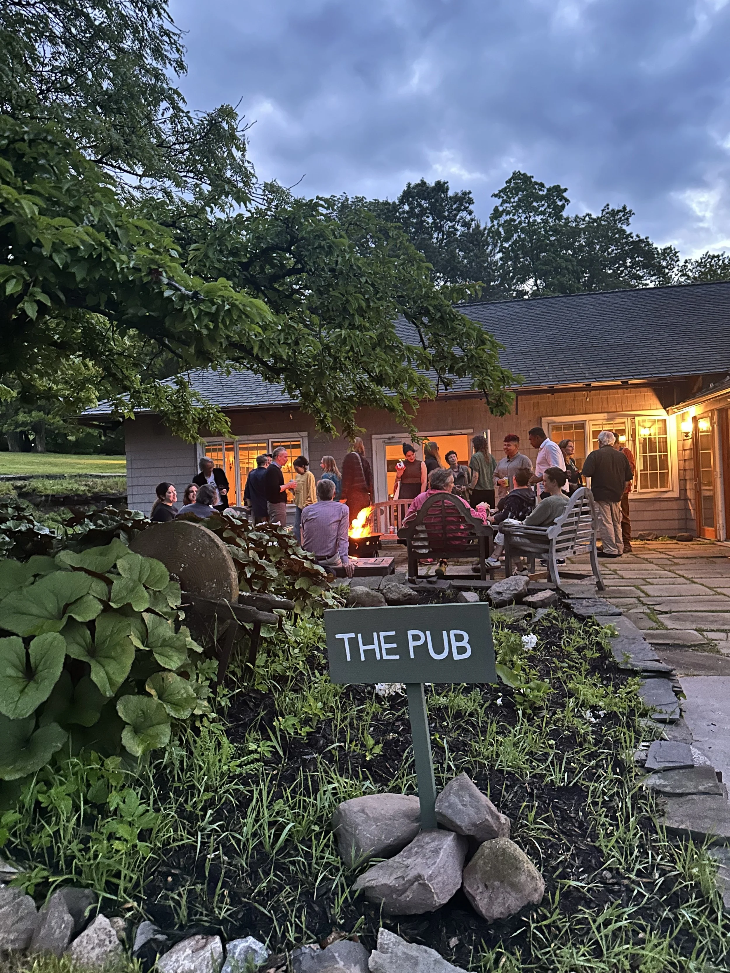 People gathered around a fire pit outside a house, with a sign in the foreground reading 'THE PUB', in a garden during evening time.
