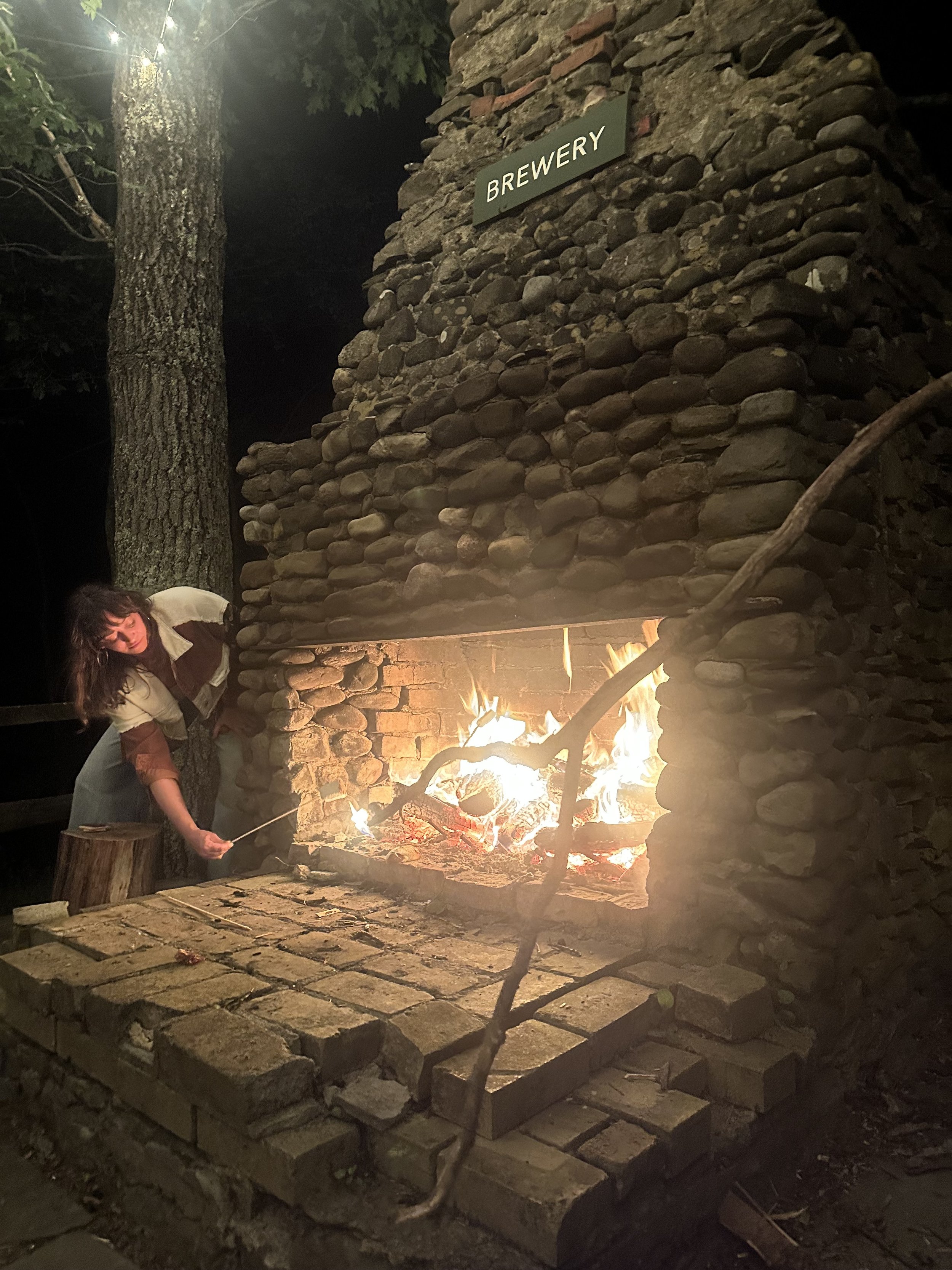 A woman standing next to a brick outdoor fireplace or fire pit, tending a fire with a long stick, under a wooden and stone structure labeled 'BREWERY' at night.