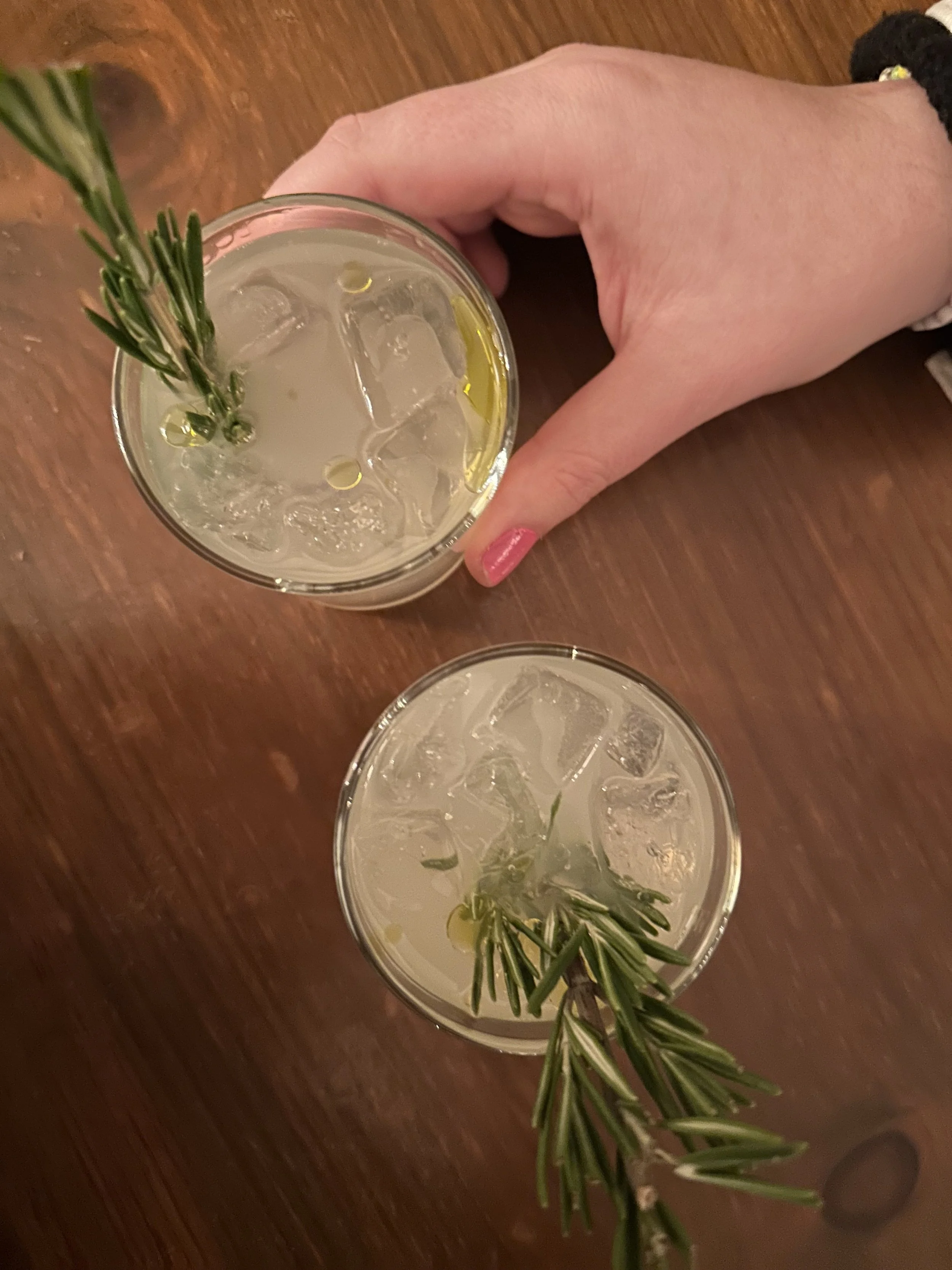 Two glasses of lemonade garnished with sprigs of fresh rosemary and ice cubes on a wooden table, viewed from above.