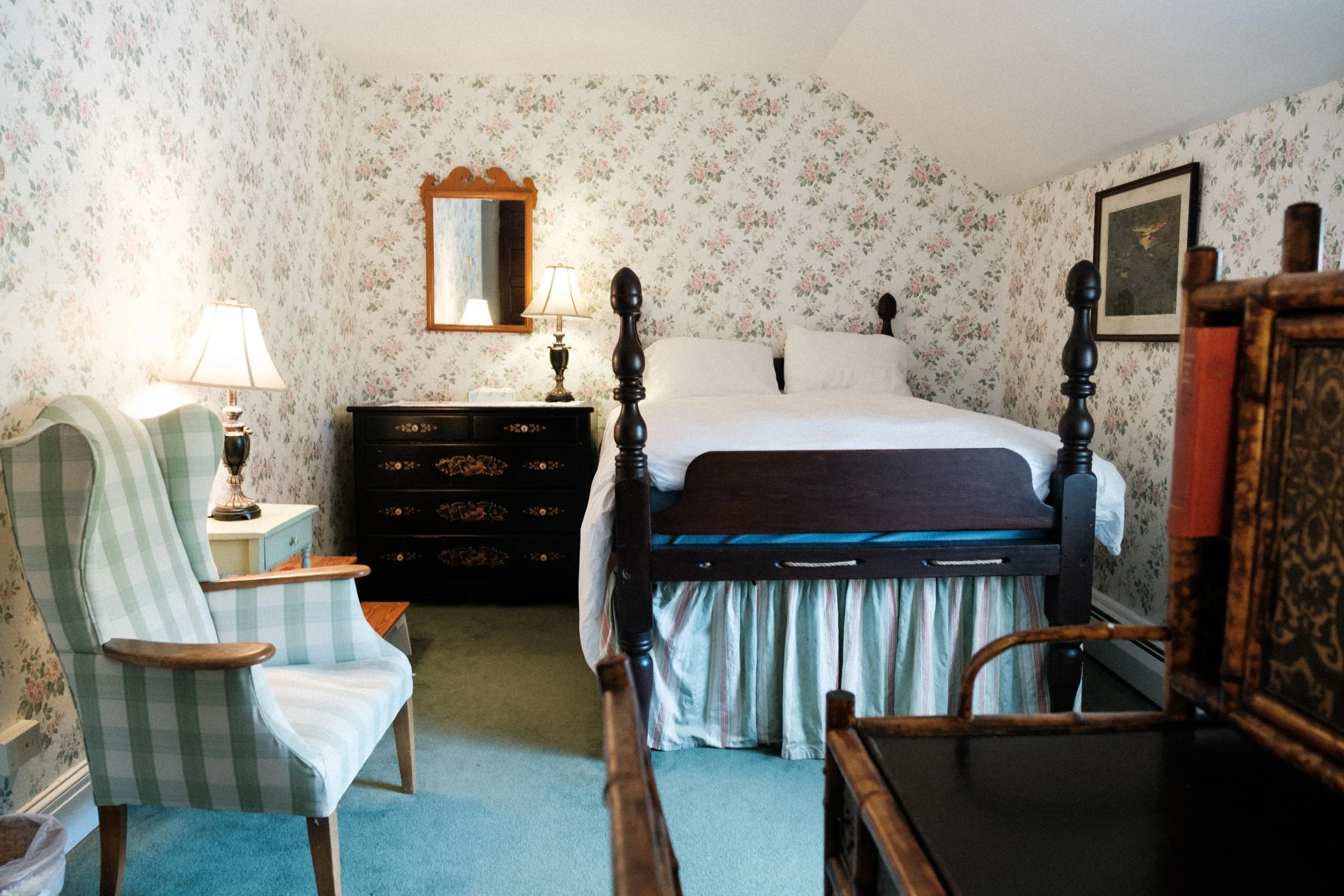 A vintage-style bedroom with floral wallpaper, a four-poster bed with white bedding and a striped bed skirt, a black dresser with decorative gold accents, two matching lamps, a framed mirror, and an armchair with green and white checkered upholstery.
