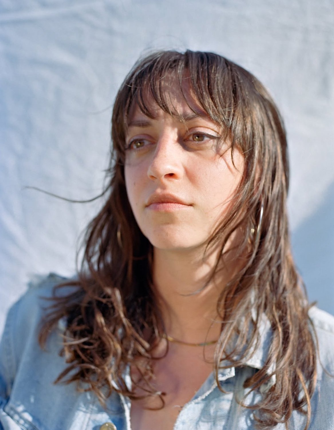 Close-up of a young woman with shoulder-length brown hair, wearing hoop earrings and a denim jacket, looking off to the side with a neutral expression, standing outdoors against a light-colored background.