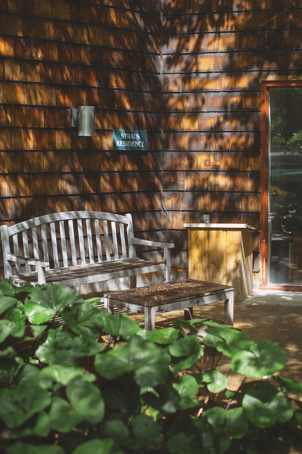 A wooden bench and table on a patio outside a building with wooden shingles. There is a small green sign on the wall that says "Straus Residence." Shadows of tree branches are cast on the building. Green leaves are in the foreground.