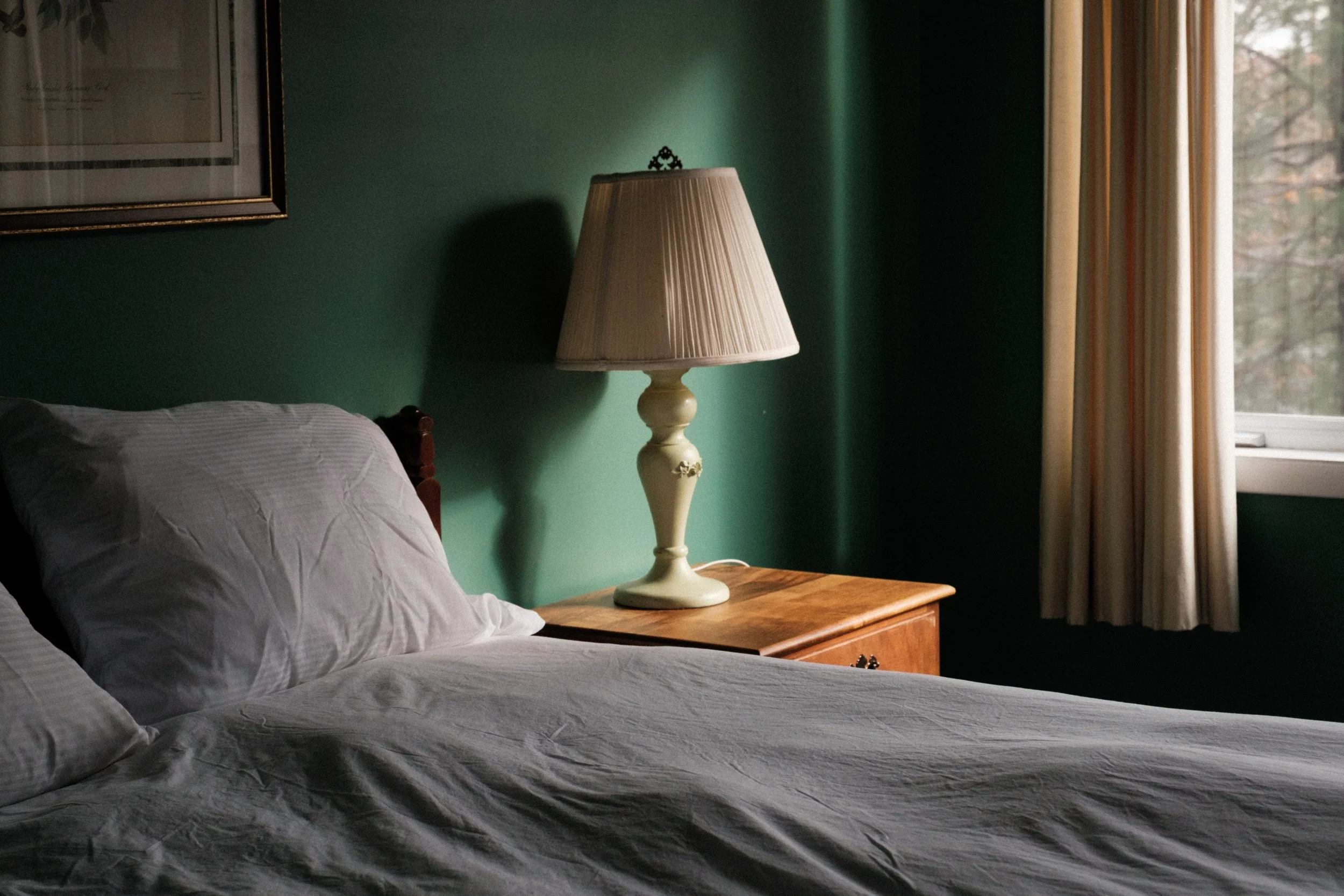 A bed with white linen on a wooden nightstand, a table lamp, a green wall, a window with cream curtains, and a framed picture on the wall.