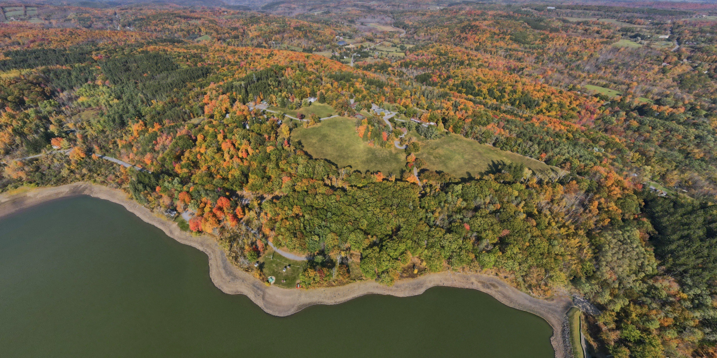 An aerial view of a lush landscape with a lake along the shoreline, surrounded by a dense forest with colorful autumn foliage and open grassy areas.