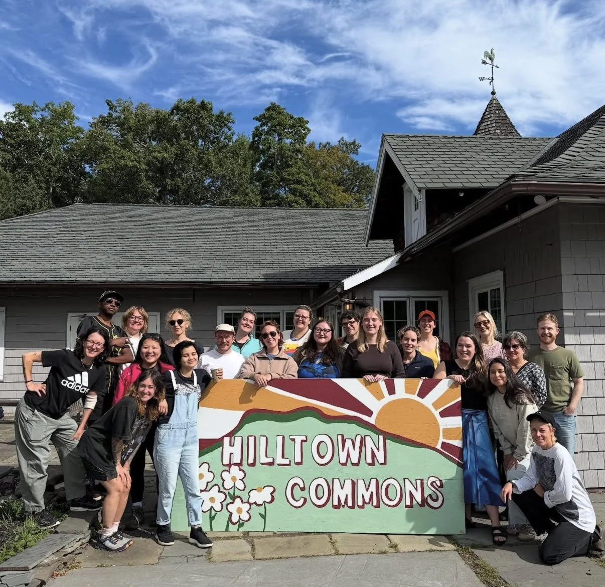 A group of people standing outdoors in front of a house, holding a colorful sign that reads 'HILLTOWN COMMONS' with a sun, mountains, and flowers painted on it, under a blue sky with clouds.