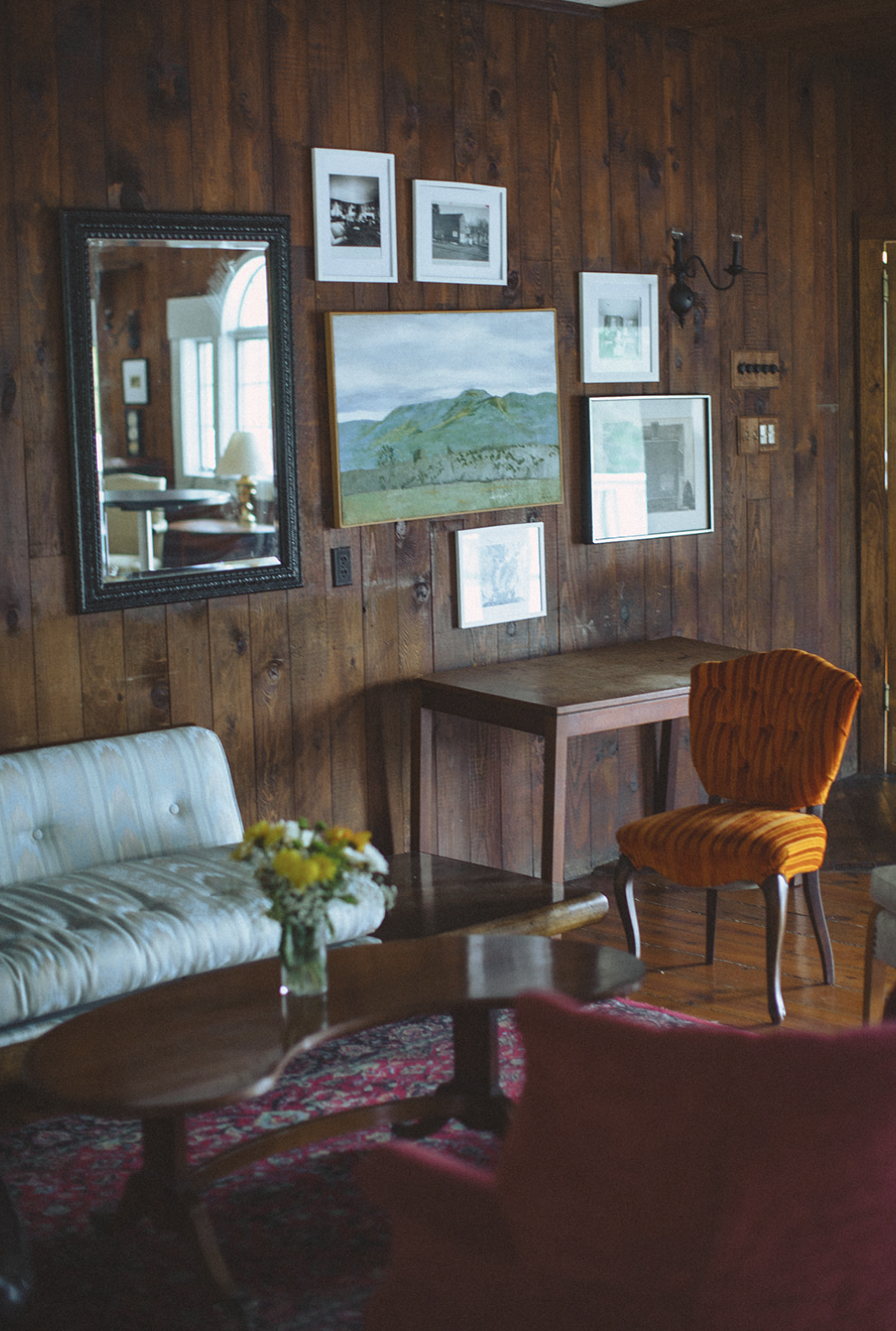 A living room with wooden walls decorated with framed photographs and paintings. There is a mirror on the wall reflecting a window, a vintage sofa with striped upholstery, a small wooden table with a flower vase, and an orange upholstered chair.