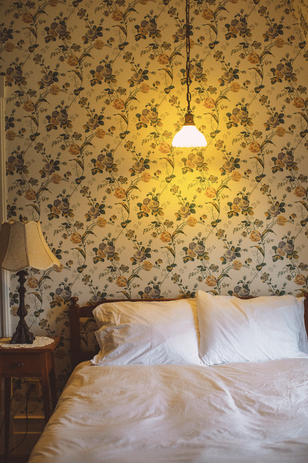A bedroom with a floral wallpaper, a bed with white pillows and a pinkish bedspread, a vintage lamp on a wooden nightstand, and a hanging ceiling light.