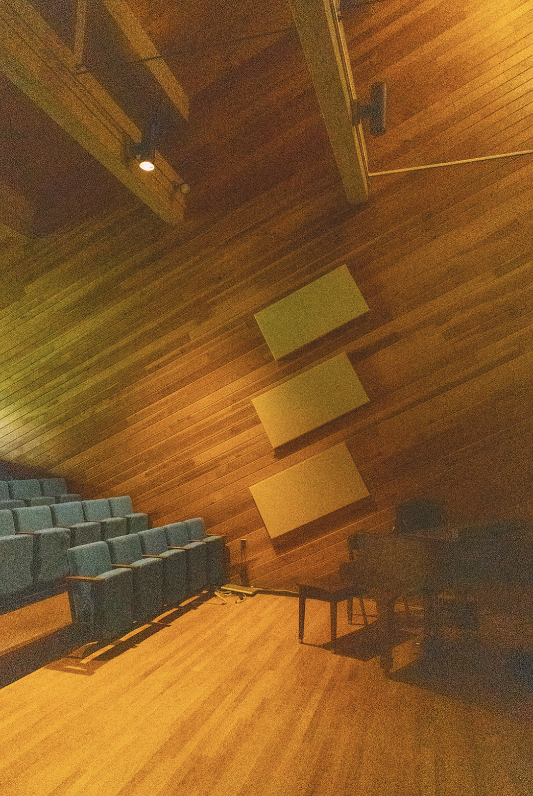 Empty auditorium with wooden walls and floor, gray seats on the left, a black Grand piano on the right, a small table, and yellow acoustic panels on the wall.