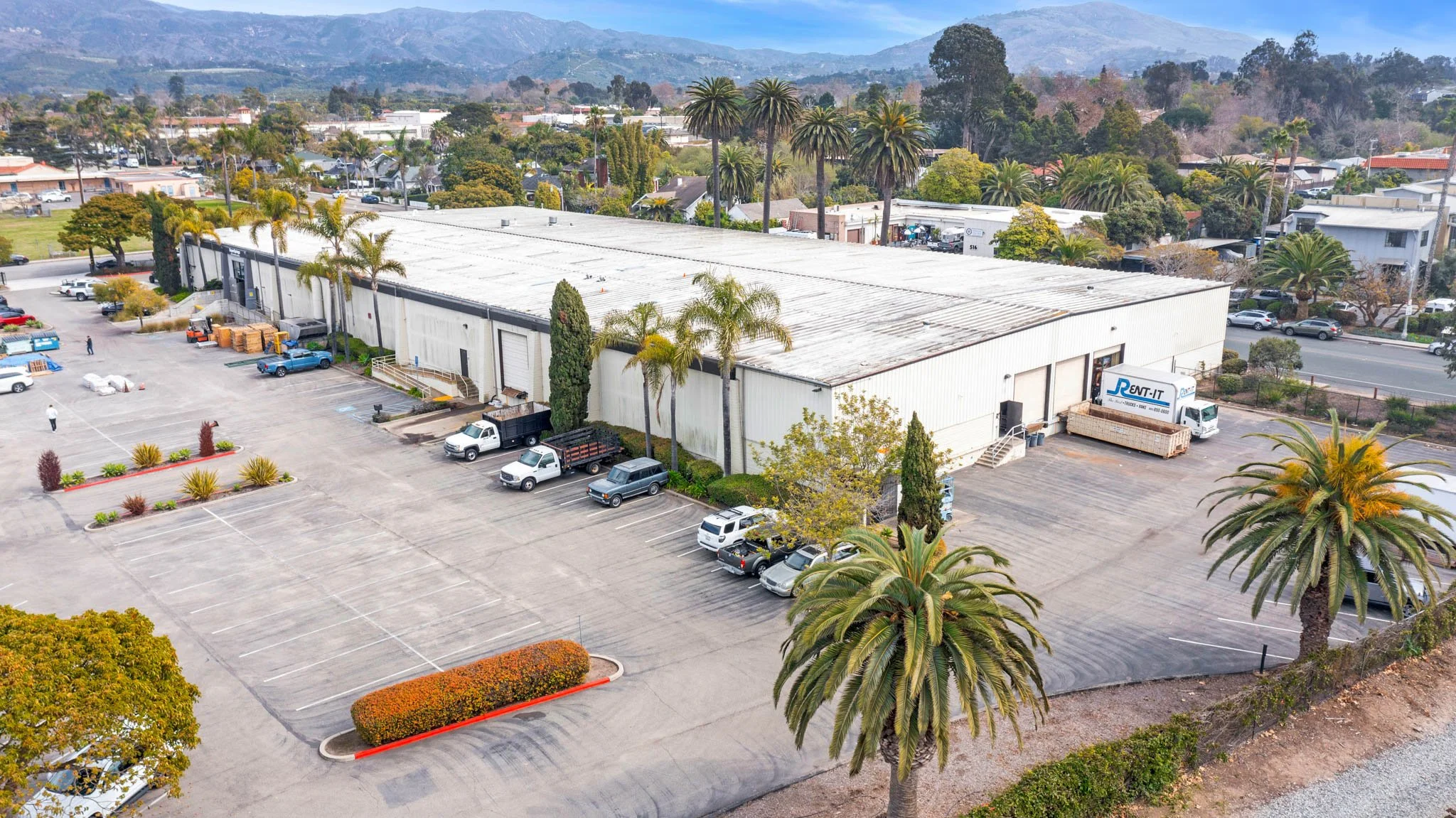 An aerial view of a commercial building with a parking lot, surrounded by palm trees and a residential neighborhood with mountains in the background.