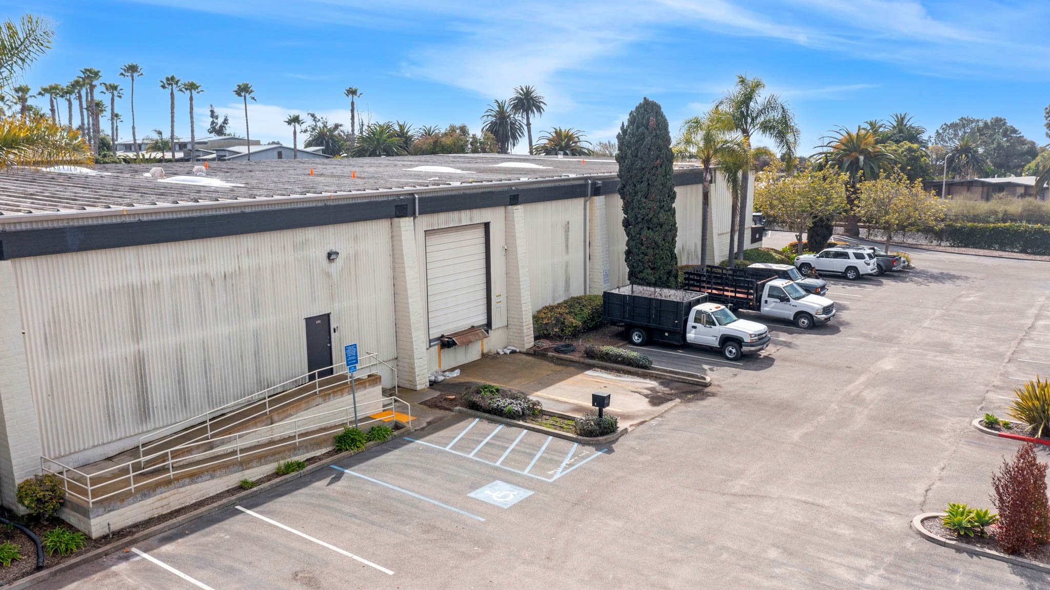 Empty parking lot with a handicapped parking space, a loading dock with a ramp, and several white trucks parked near a beige industrial building, surrounded by palm trees and bushes.