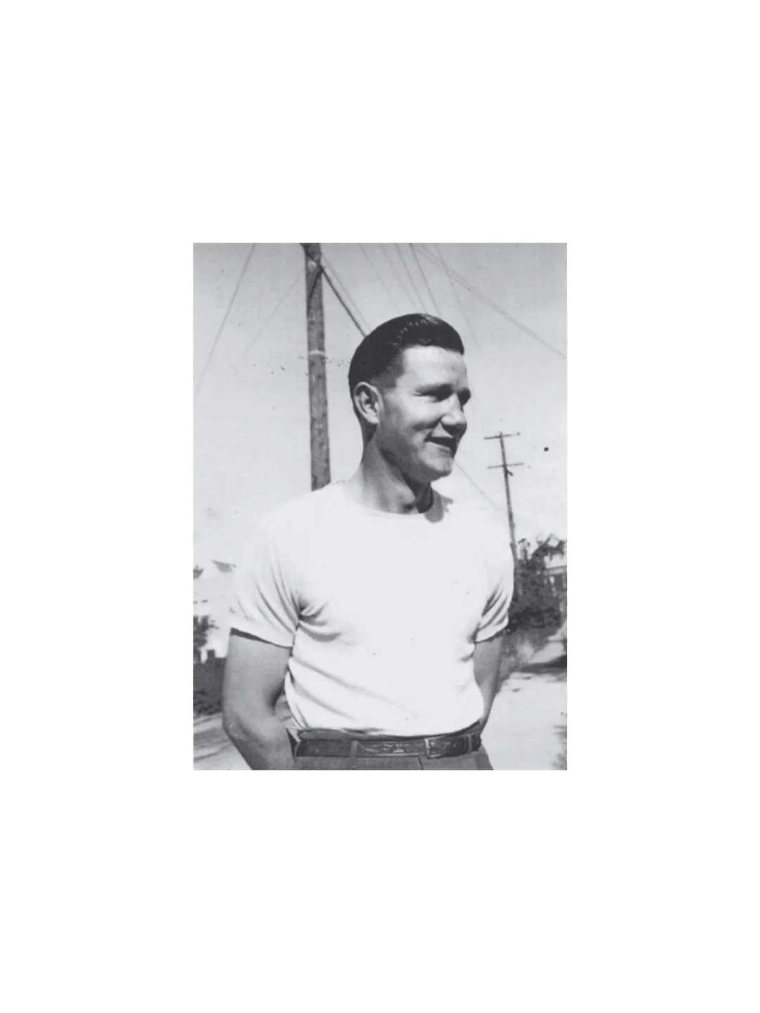 A black and white photo of a smiling young man with slicked-back hair, wearing a white T-shirt and dark trousers, standing outdoors with power lines and utility poles in the background.