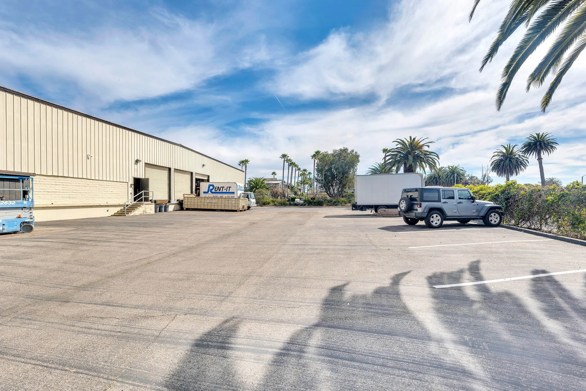 Empty parking lot outside a warehouse with a few parked vehicles, palm trees, and a partly cloudy sky.