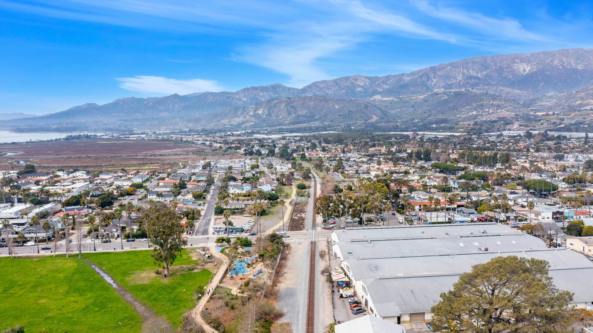 Aerial view of a suburban area with houses, roads, greenery, a playground, train tracks, and mountains in the background under a partly cloudy sky.