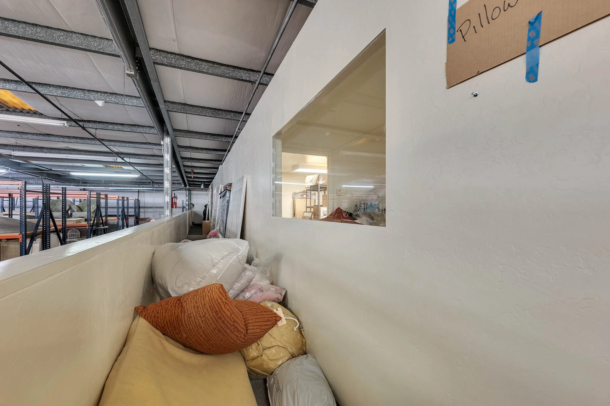 A warehouse or storage space with pillows and cushions piled along a beige wall, including a visible window into an adjacent room with shelves and equipment.