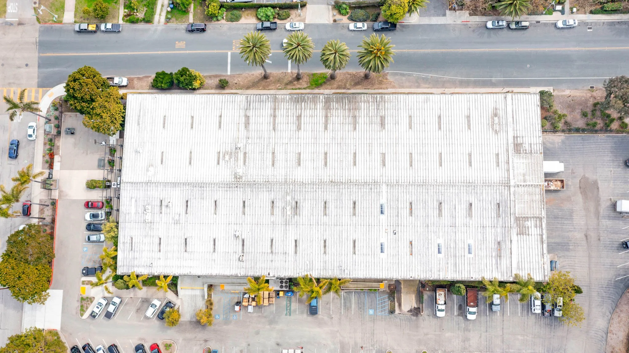 An aerial view of a large commercial building with a white roof, surrounded by parking lots with cars and trees, including palm trees along the street.