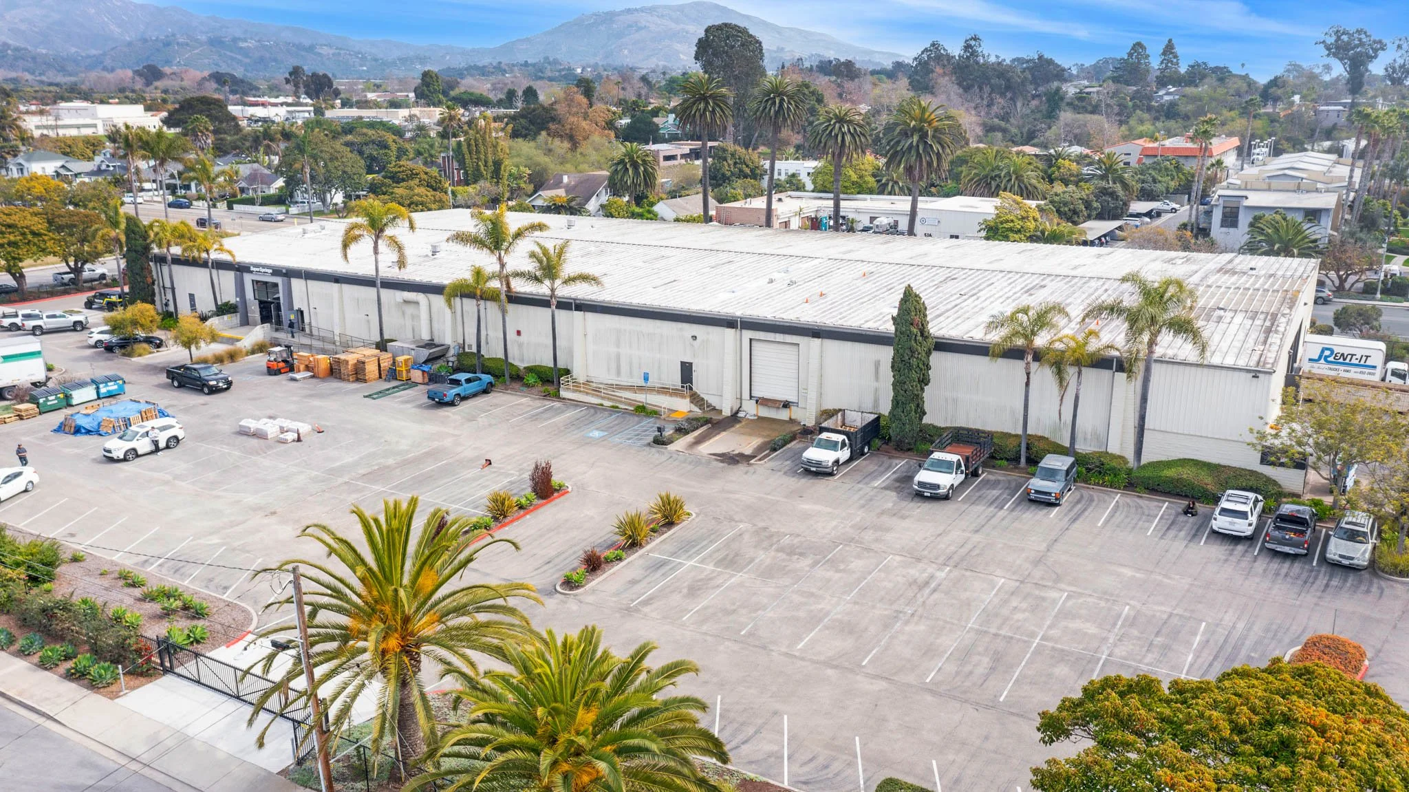 An industrial warehouse with trees and parked cars in a lot, set in a suburban area with mountains in the background.
