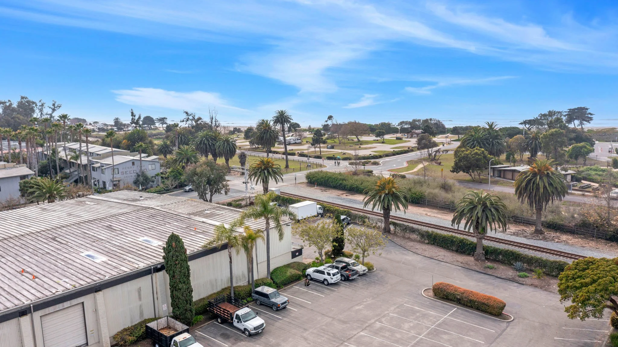 A view of a parking lot with several parked cars, adjacent to a industrial building, with palm trees and greenery, and train tracks running through a landscaped park with walking paths and trees under a partly cloudy blue sky.