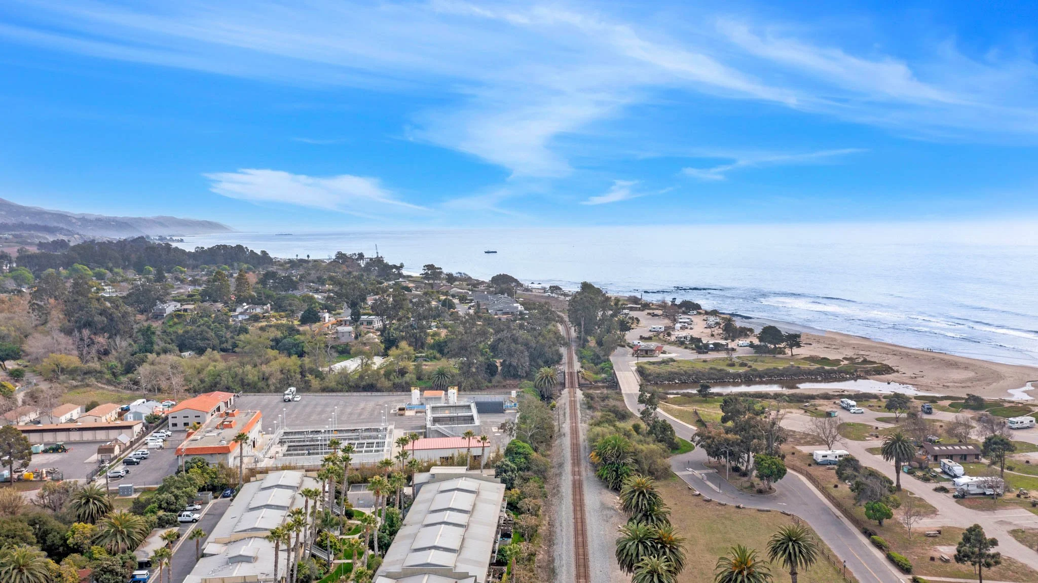Aerial view of a coastal town with houses, trees, a beach, and the ocean under a blue sky with scattered clouds.