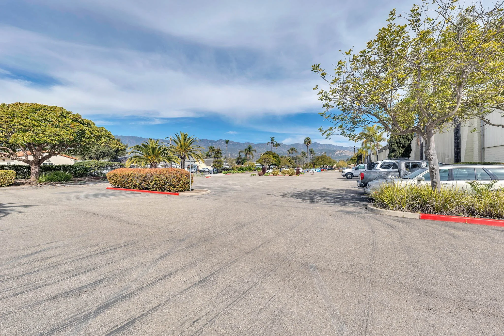 Empty parking lot with trees and mountains in the background under partly cloudy sky.