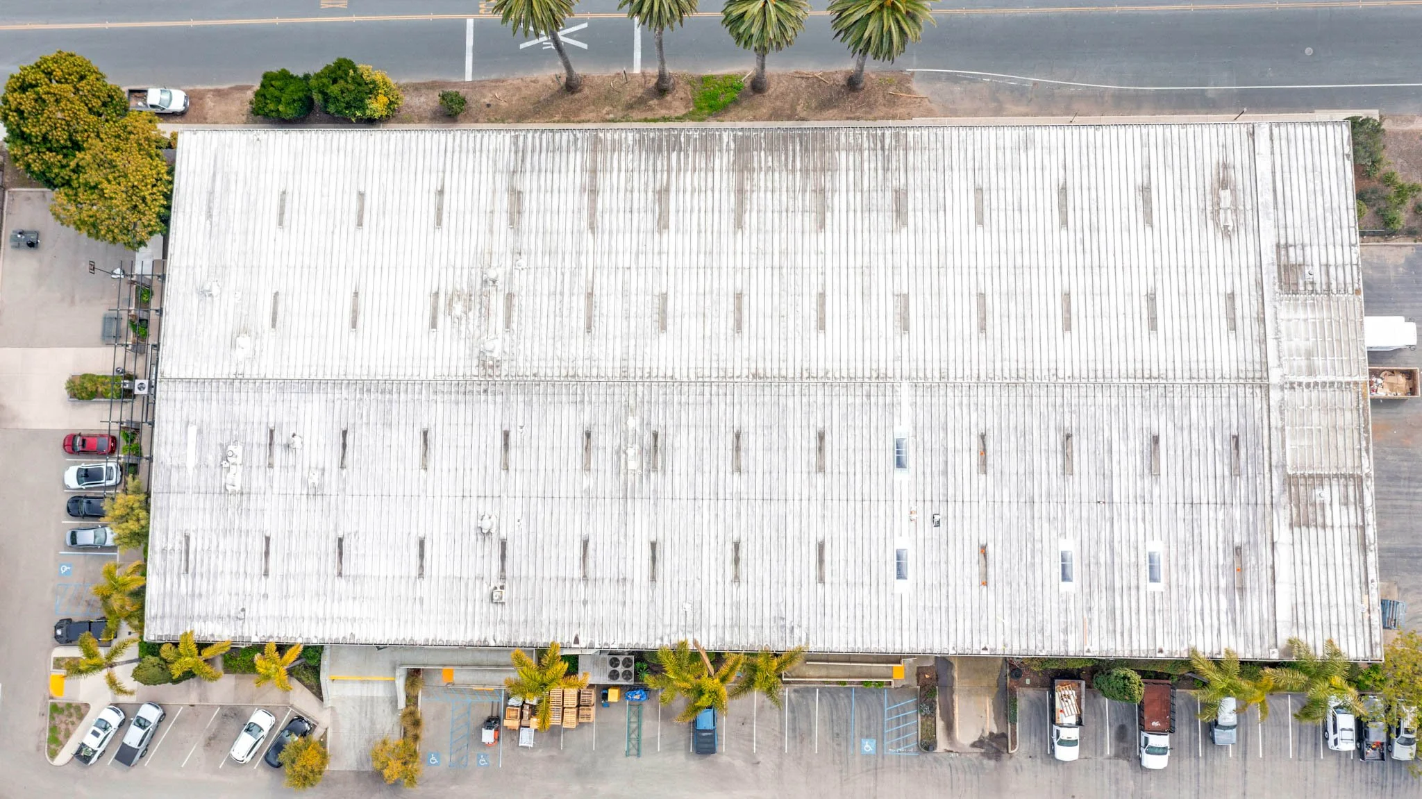 An aerial view of a large, flat-roofed commercial building with parked cars around it, some trees, and a street at the top.