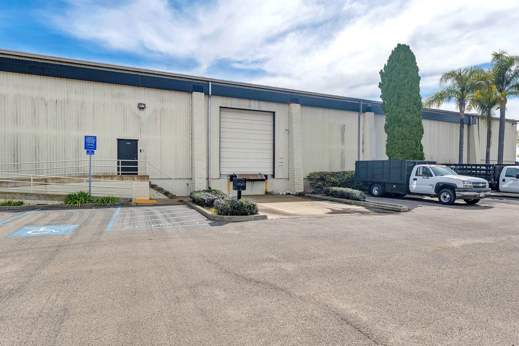 Empty parking lot in front of a warehouse with loading dock, a wheelchair parking space, and a truck parked nearby.