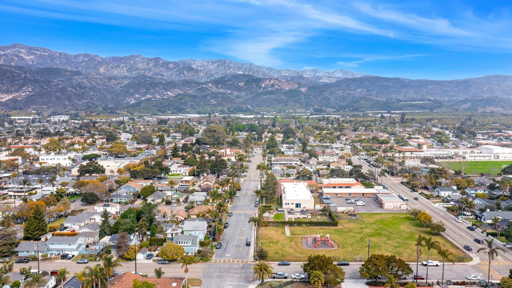 Aerial view of a small town with mountains in the background, streets lined with trees, a playground in a grassy park, and various residential and commercial buildings.