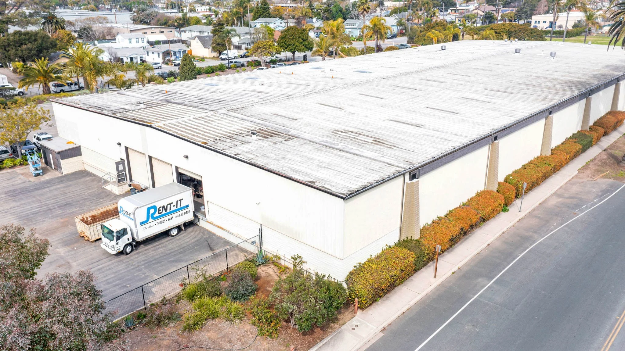Aerial view of a large commercial warehouse with a weathered roof, parking lot, and a moving truck labeled "RENT-IT" parked near the entrance. Surrounding areas include trees, small shrubs, residential houses, and palm trees.