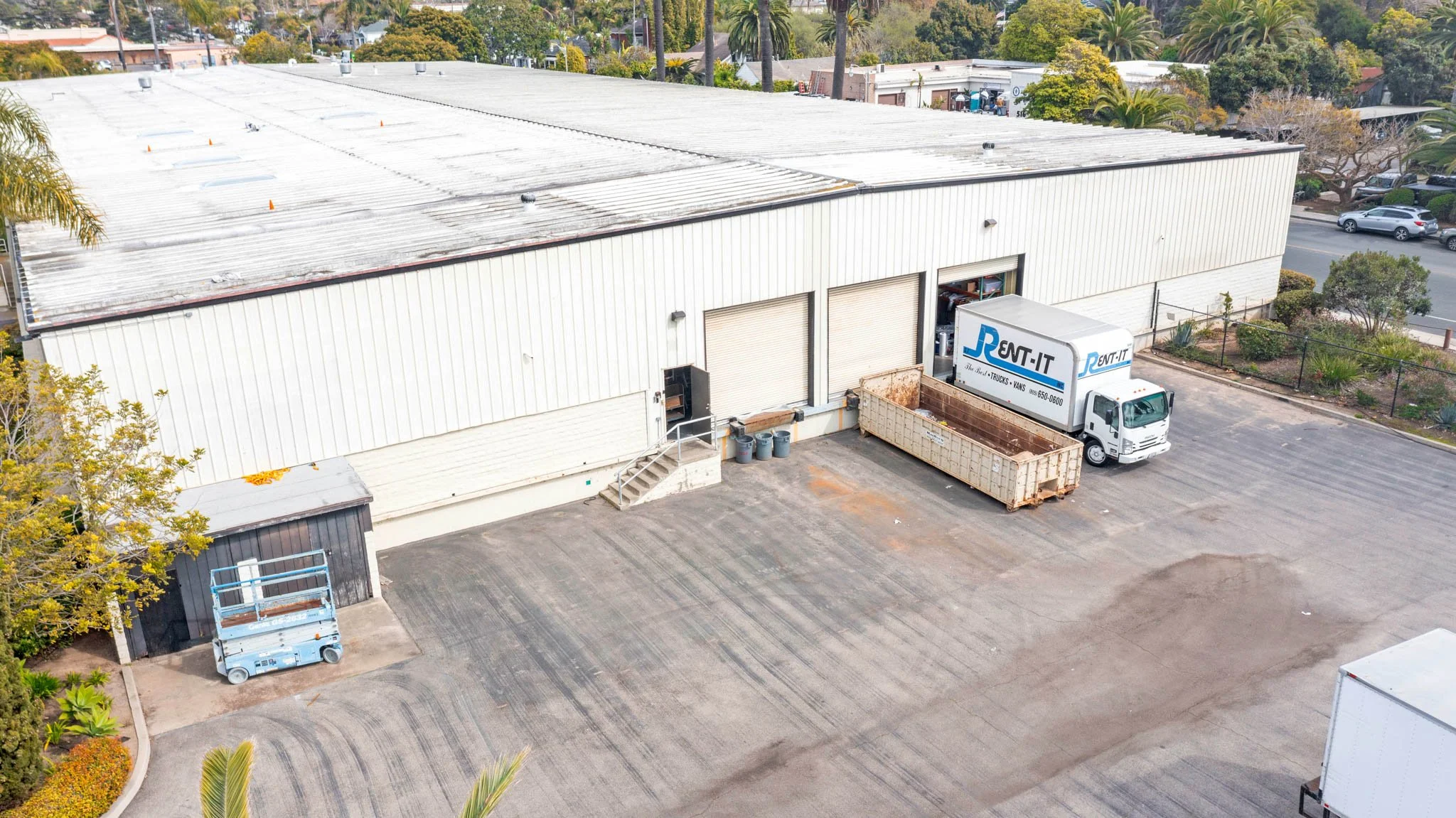 Aerial view of a warehouse building with a parking lot and delivery trucks outside. The warehouse has three large garage doors, with one open, showing a truck delivering. There is a small black shed, a scissor lift, and some trees and cars visible ar