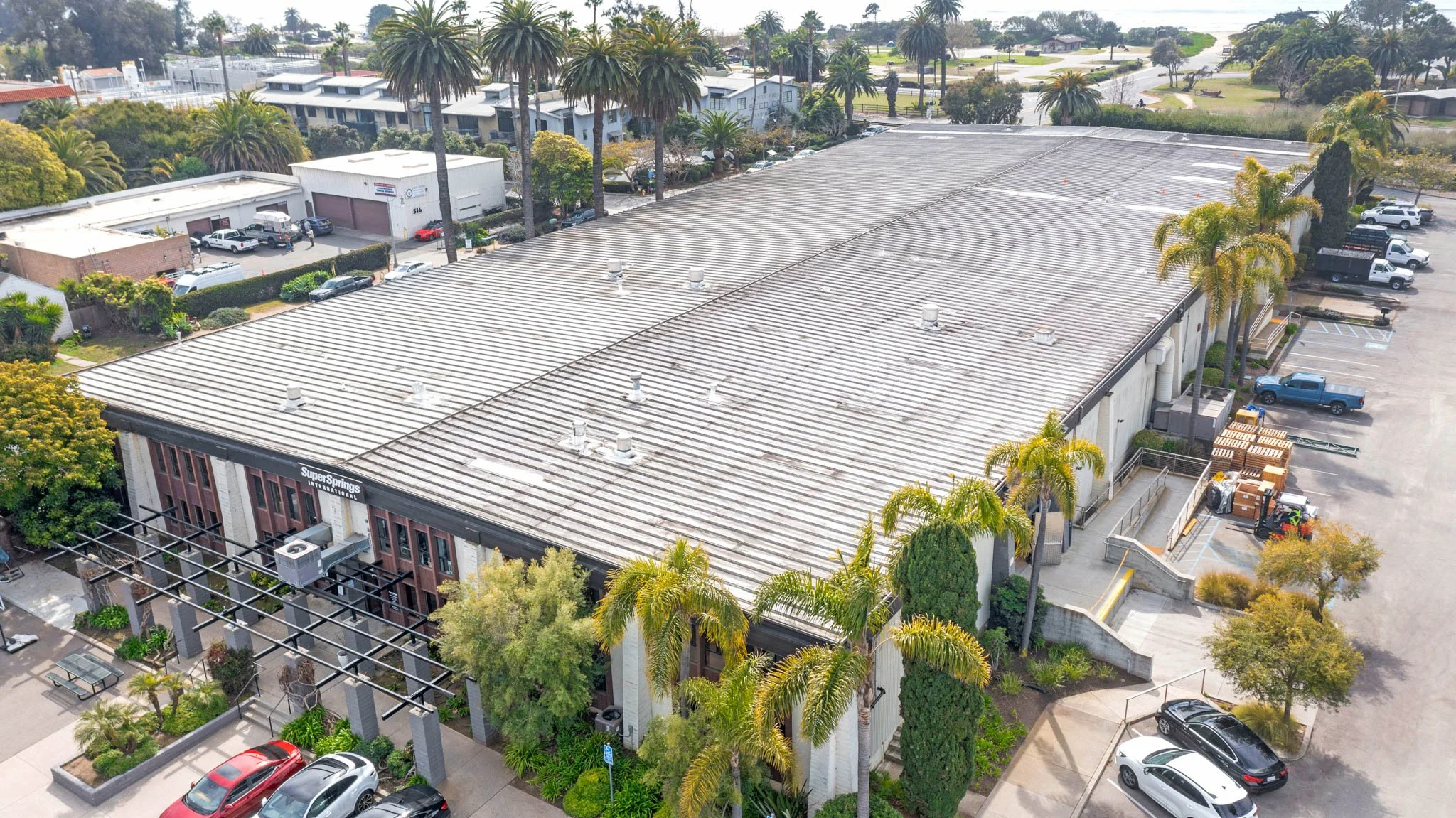 Aerial view of a commercial building with a gray metal roof, palm trees surrounding the building, and a parking lot with several cars nearby.