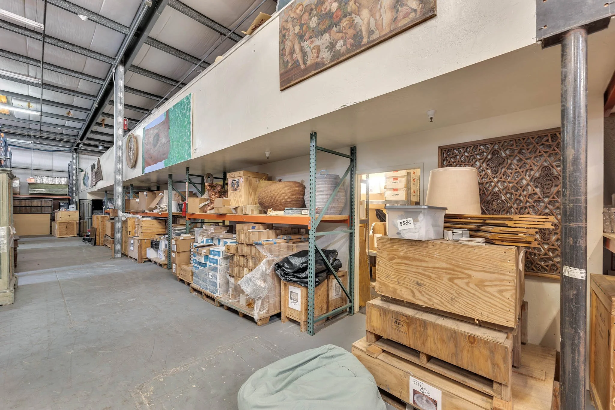 Interior of a warehouse or storage room with metal shelving, wooden crates, boxes, and various household items, including a lamp and decorative wooden panels.