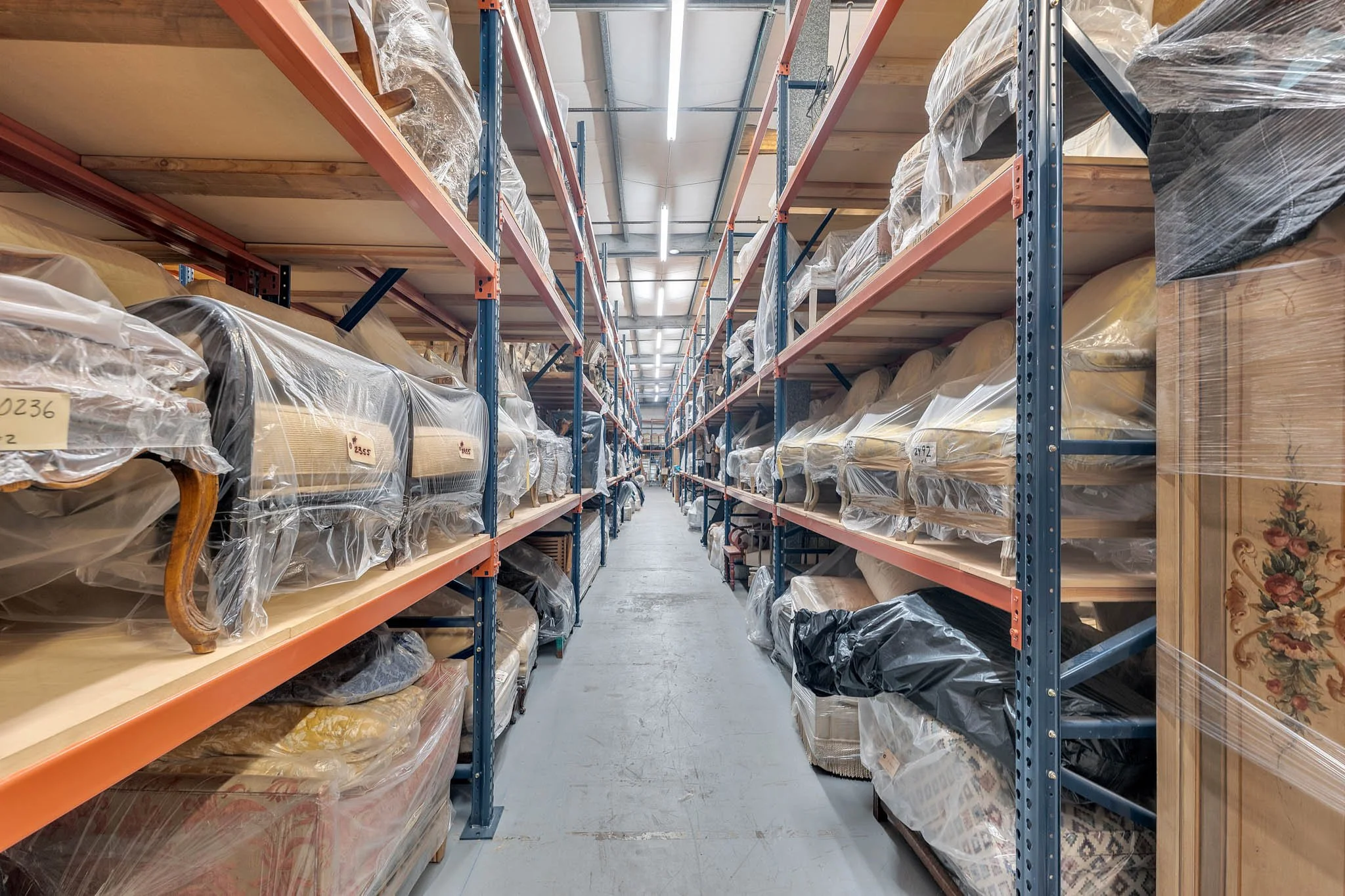 Warehouse aisle with metal shelving units holding wrapped furniture and rugs, with a concrete floor and fluorescent lighting.