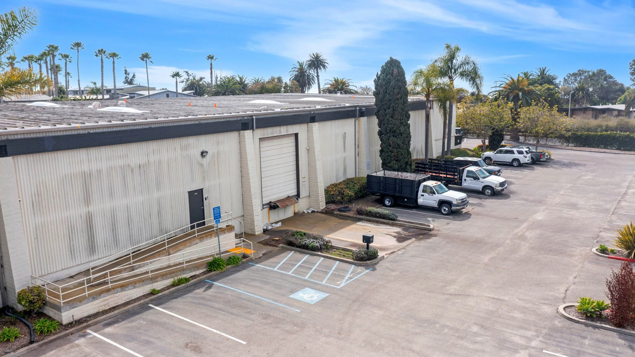 Empty parking lot with several parked trucks near a large industrial building under a clear blue sky with palm trees in the background.