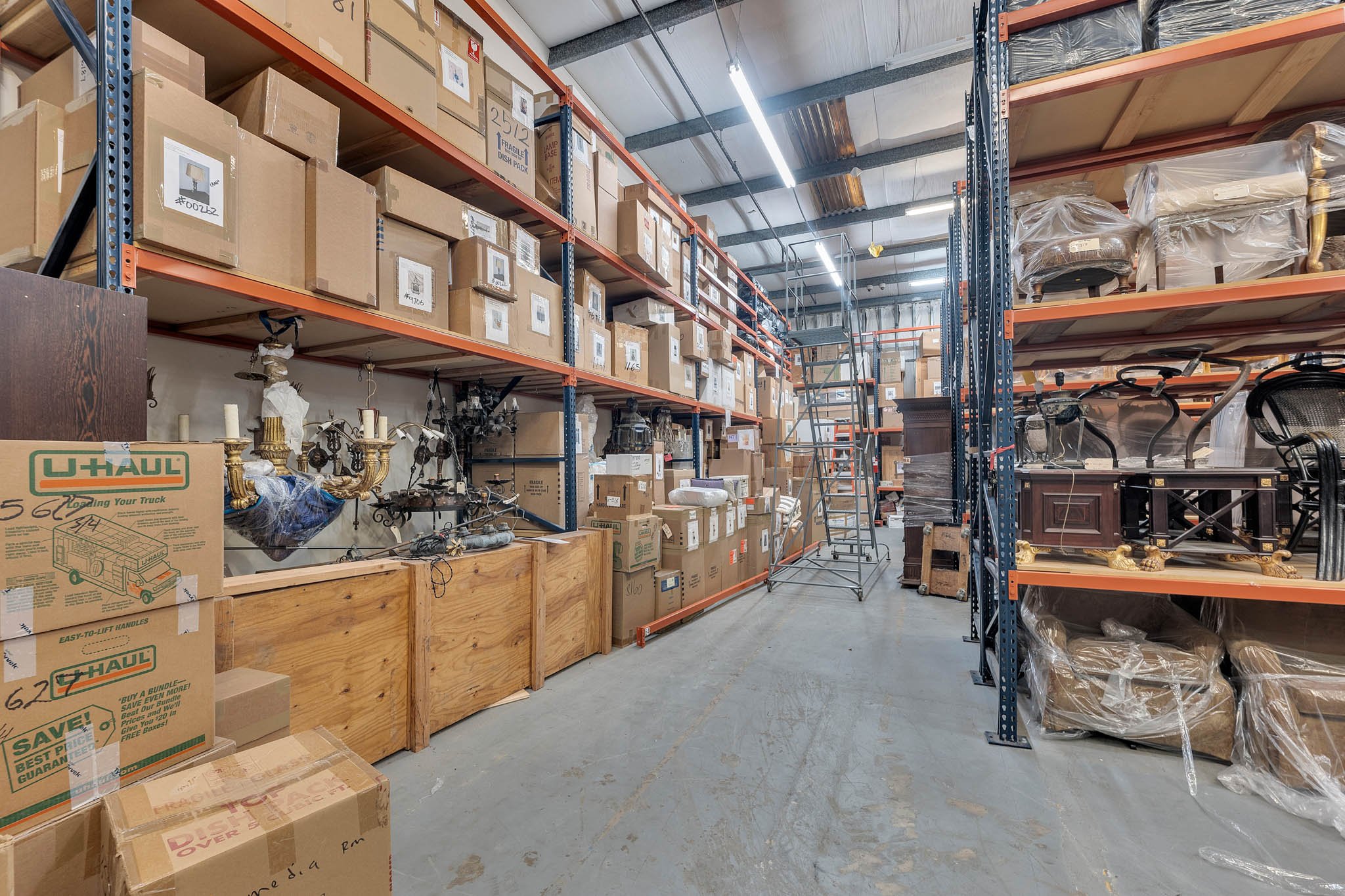 Interior view of a warehouse storage area with metal shelving filled with stacked cardboard boxes, some wrapped furniture pieces, ladders, and various antique light fixtures on wooden pallets.