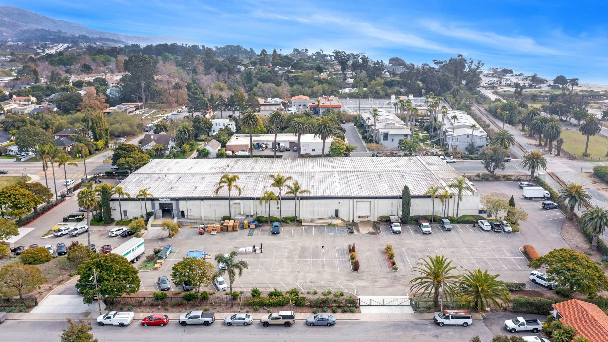 Aerial view of a commercial warehouse with a parking lot, surrounded by trees, neighboring residential houses, and a distant coastline with hills.