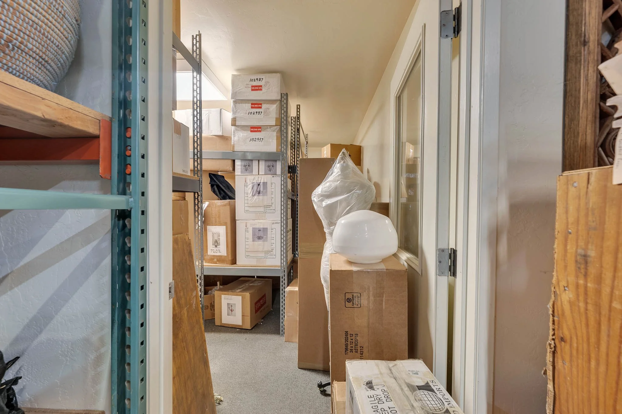 A cluttered storage closet with shelves holding boxes and various items, including a white decorative object on a cardboard box, and a large window on the right wall.