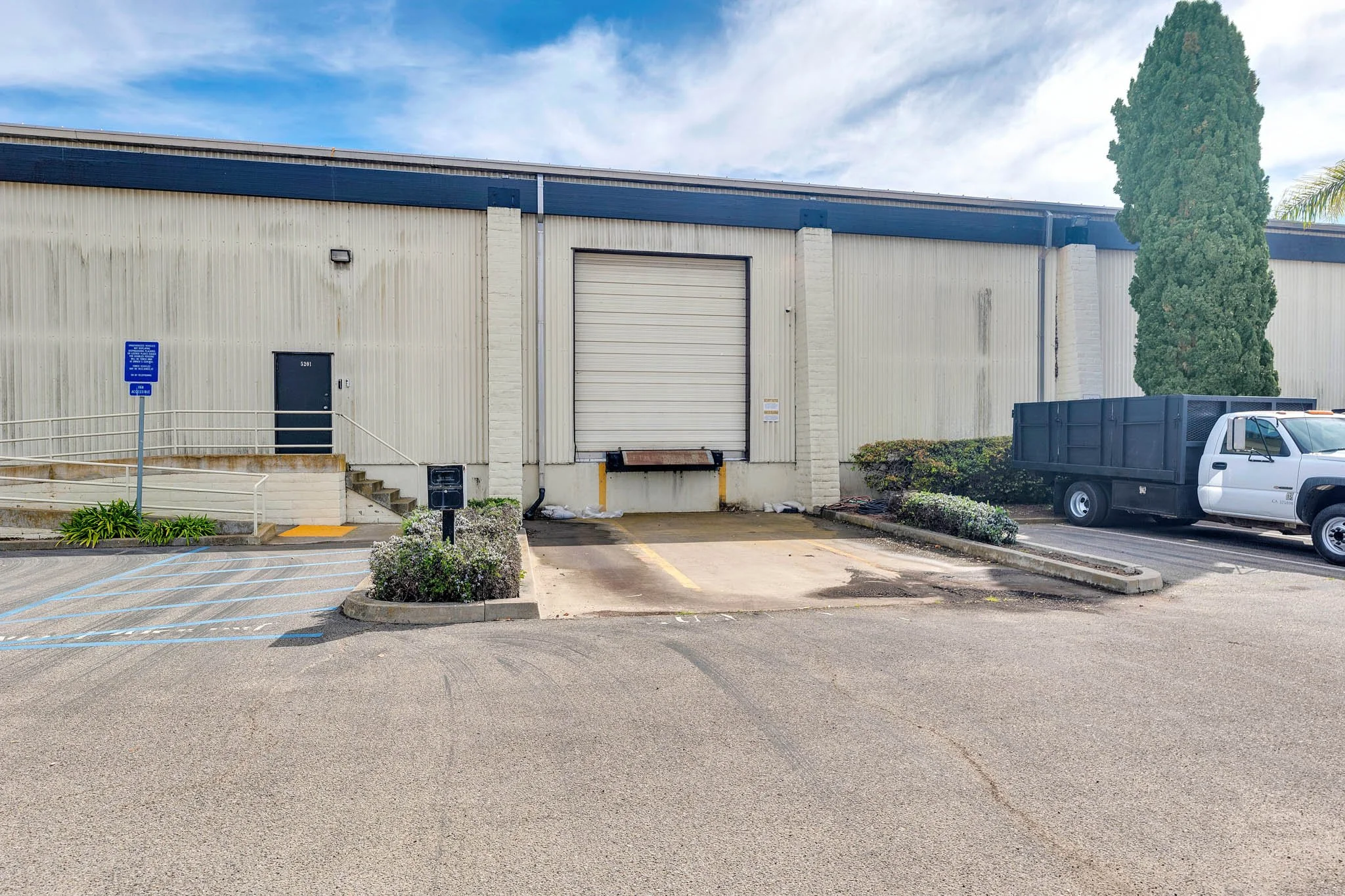 Loading dock with a closed roll-up door, adjacent to a parking space, a small bush, a tall tree, and a white truck with a black container bed in a commercial area under a partly cloudy sky.