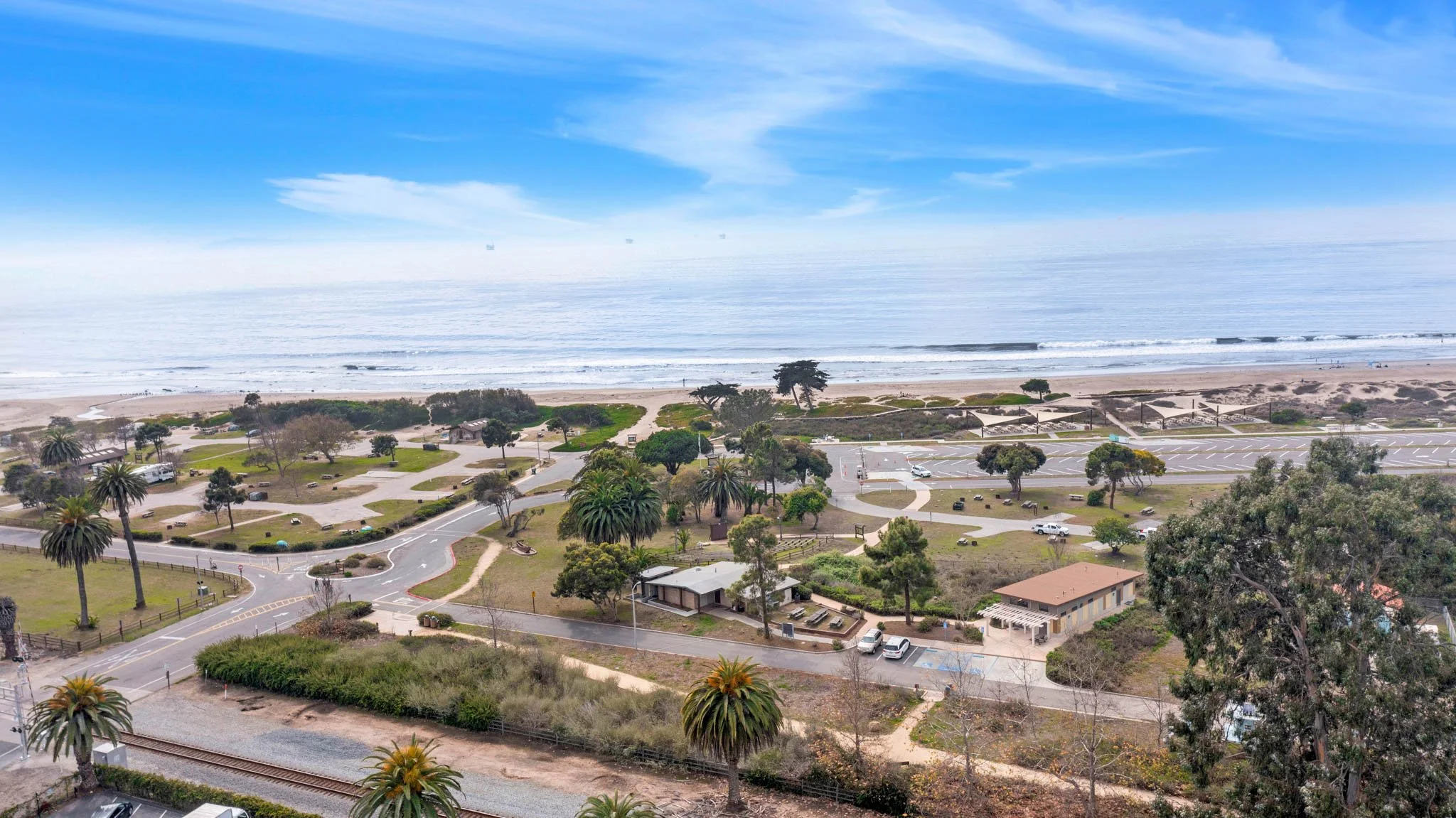 Aerial view of a coastal park with palm trees and grassy areas, adjacent to a beach and ocean with distant ships, and a parking lot along the shoreline.