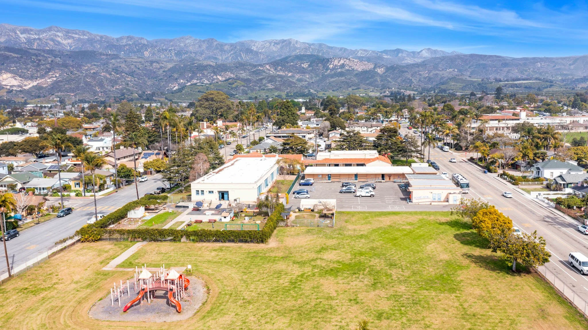 Aerial view of a neighborhood with houses, a playground, parking lot, and mountains in the background.