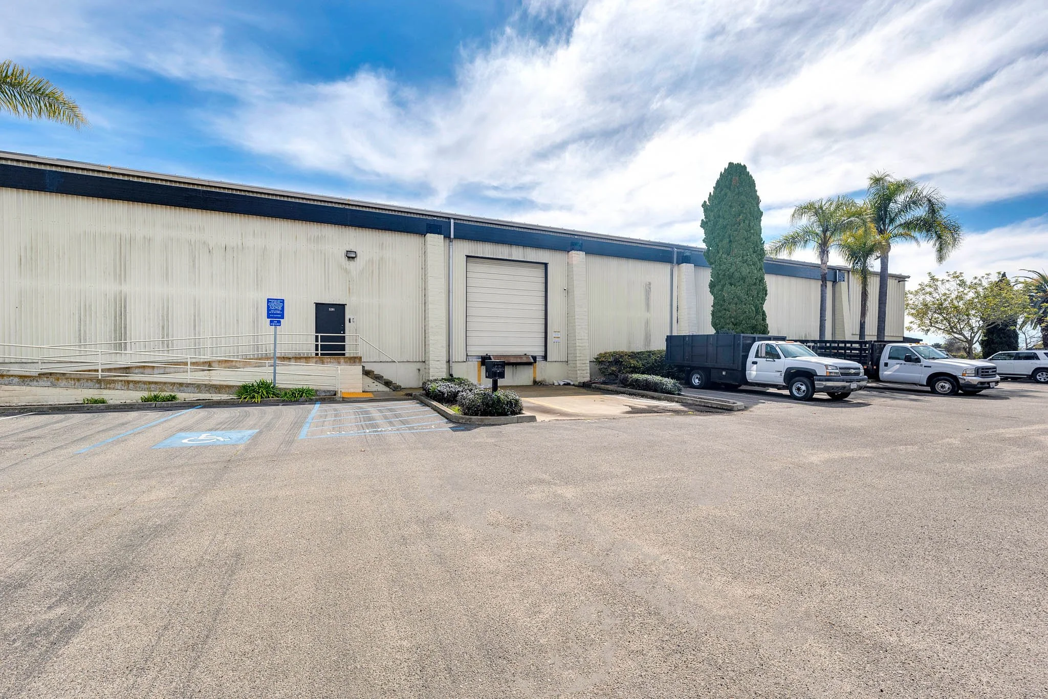 Large warehouse building with a loading dock, a designated handicapped parking space, and multiple pickup trucks parked in front. There are palm trees and a clear partly cloudy sky overhead.