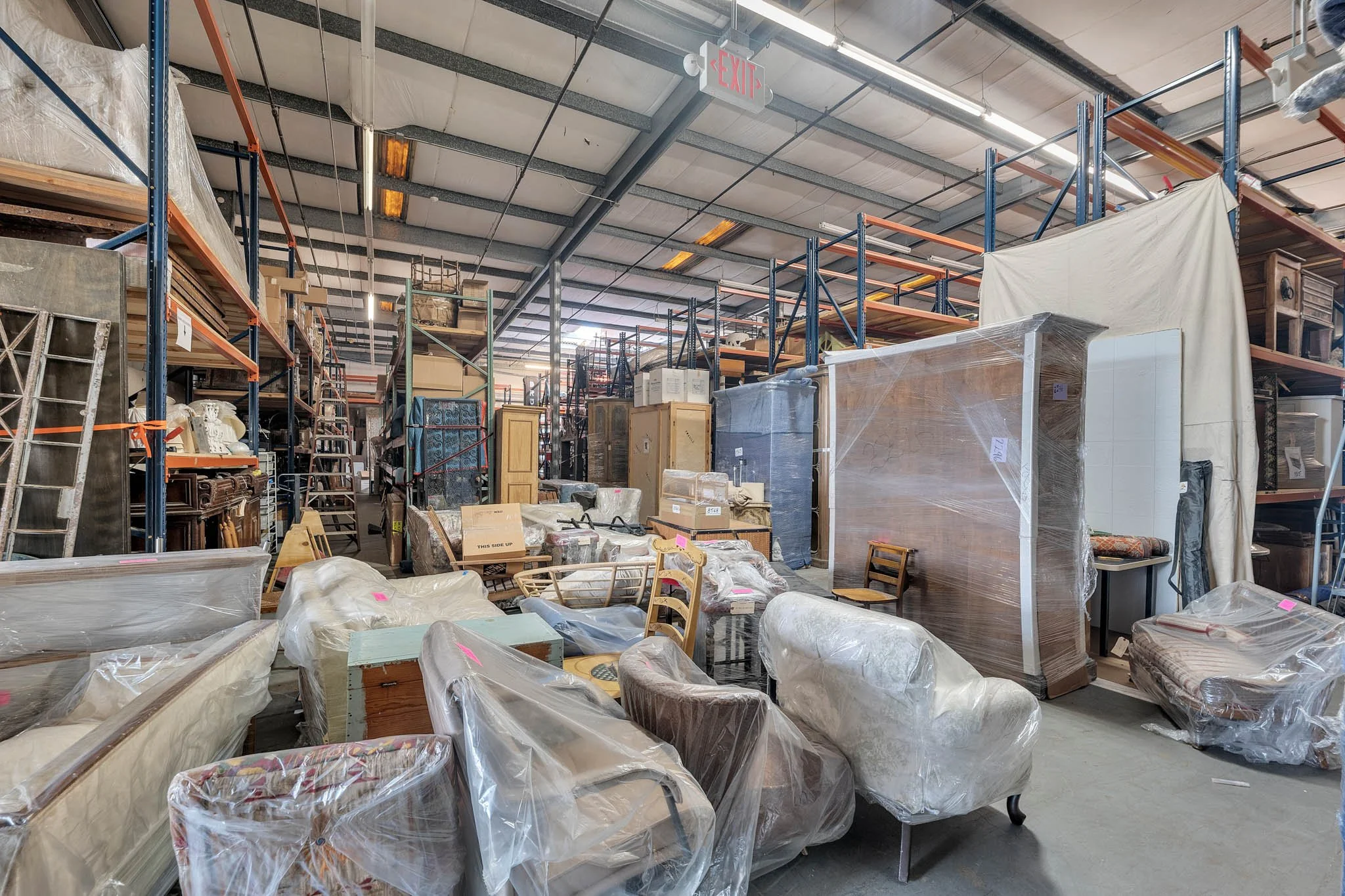 Warehouse with furniture and boxes, some wrapped in plastic, stored on shelves and stacked on the floor.