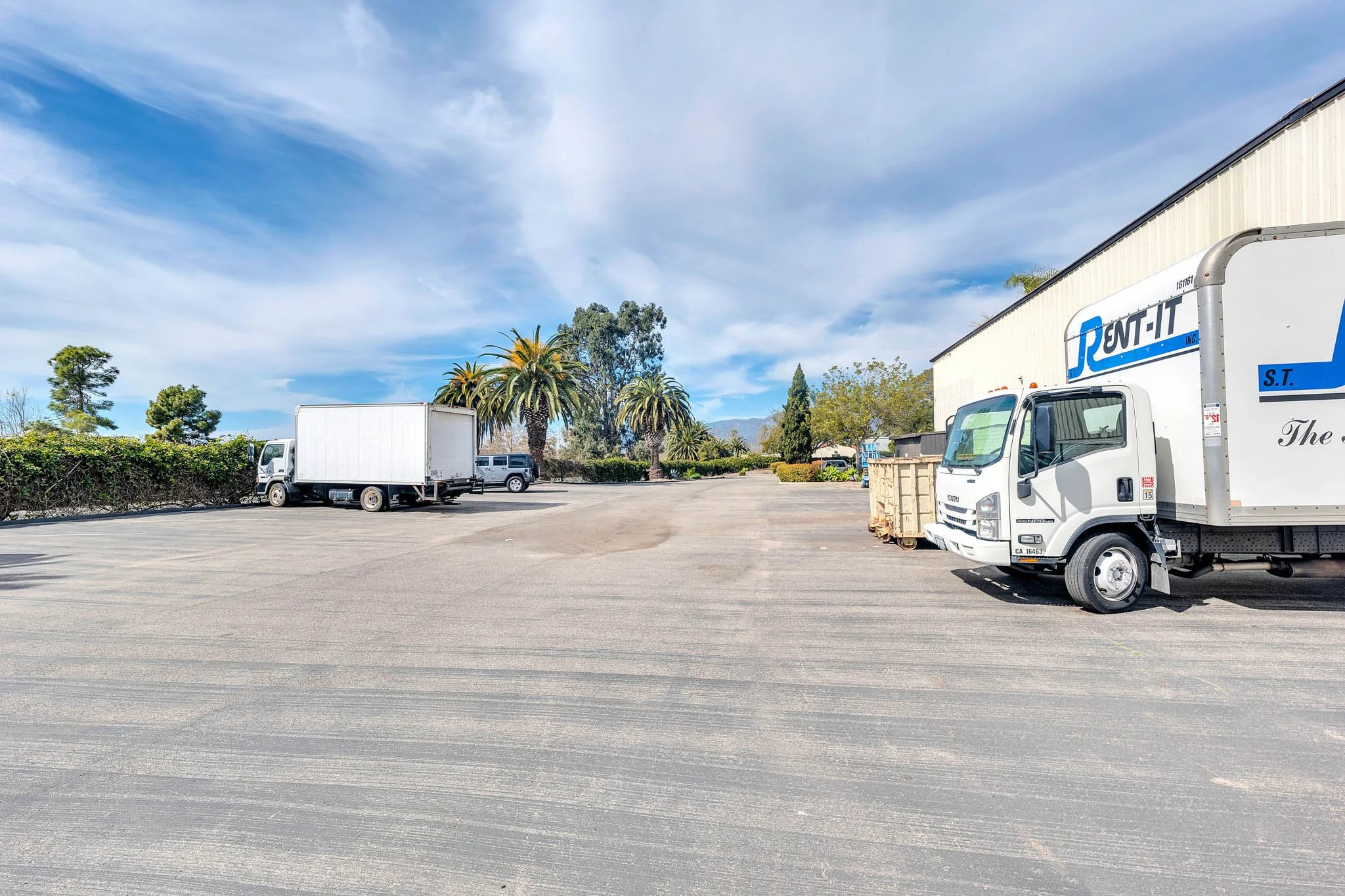An empty parking lot with a few trucks parked and palm trees in the background under a partly cloudy sky
