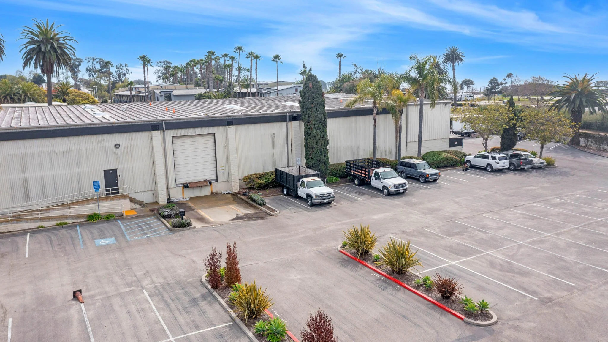 Empty parking lot with a few parked trucks near a white industrial building, surrounded by palm trees and shrubs under a blue sky.