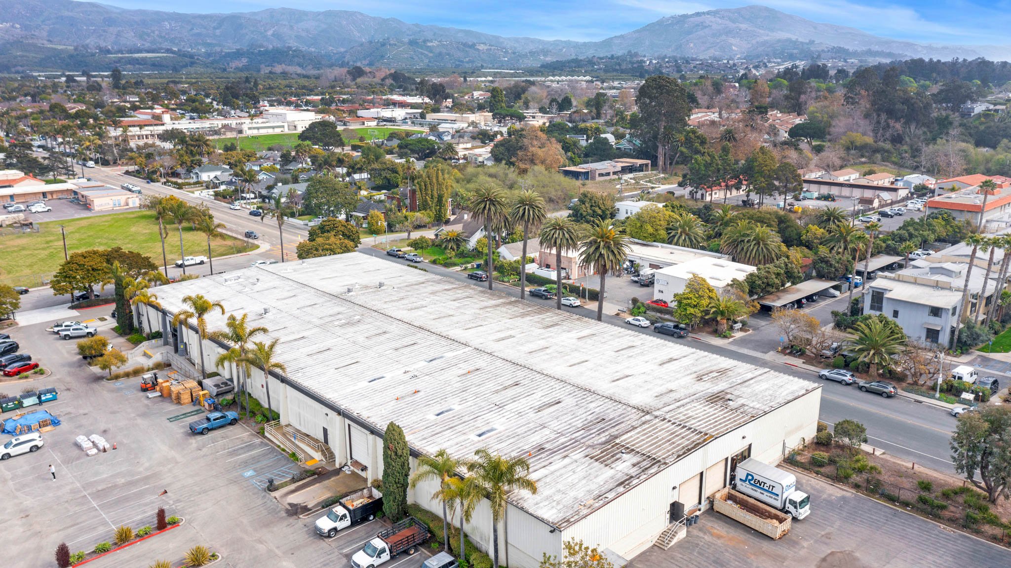 Aerial view of a commercial warehouse building with a weathered roof, surrounded by a parking lot with cars and trucks, and a nearby residential area with trees and houses, in a suburban setting with mountains in the background.